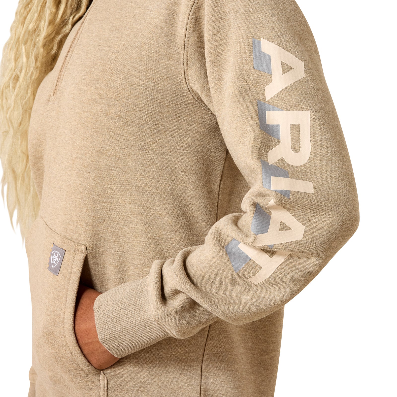 A woman wearing a light tan heathered pullover hoodie with a half-zip neckline, kangaroo pocket, and a small square logo patch near the lower front, paired with blue jeans against a plain white background.