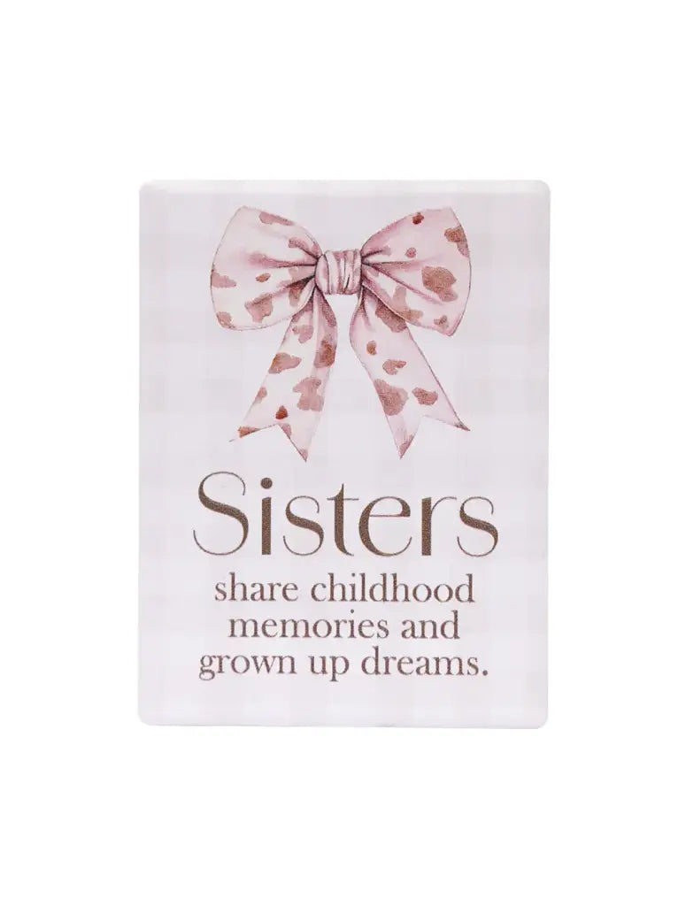 A rectangular decorative sign with rounded corners featuring a pink patterned bow at the top and the text 'Sisters share childhood memories and grown up dreams.' on a white background.