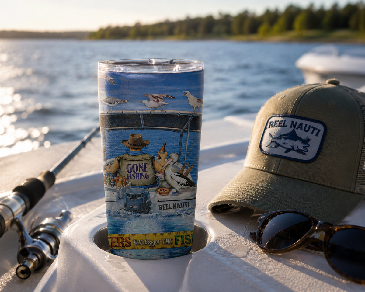 A clear insulated tumbler with a colorful fishing-themed wraparound print stands on a boat seat by the water. A tan Reel Nauti trucker hat, sunglasses, and a fishing rod are placed around it, with a lake and shoreline blurred in the background.