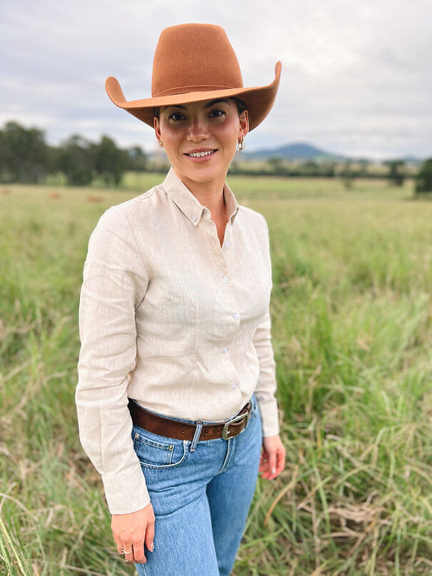 Reba Women's Stone Linen Gal L/S Shirt. A woman standing in a field wearing a stone linen long-sleeved shirt and a brown cowboy hat.