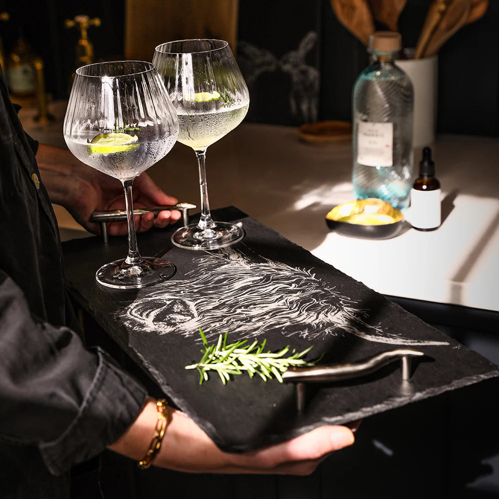 Two glasses of gin and tonic with a lemon slice on a black slate tray, held by a person.