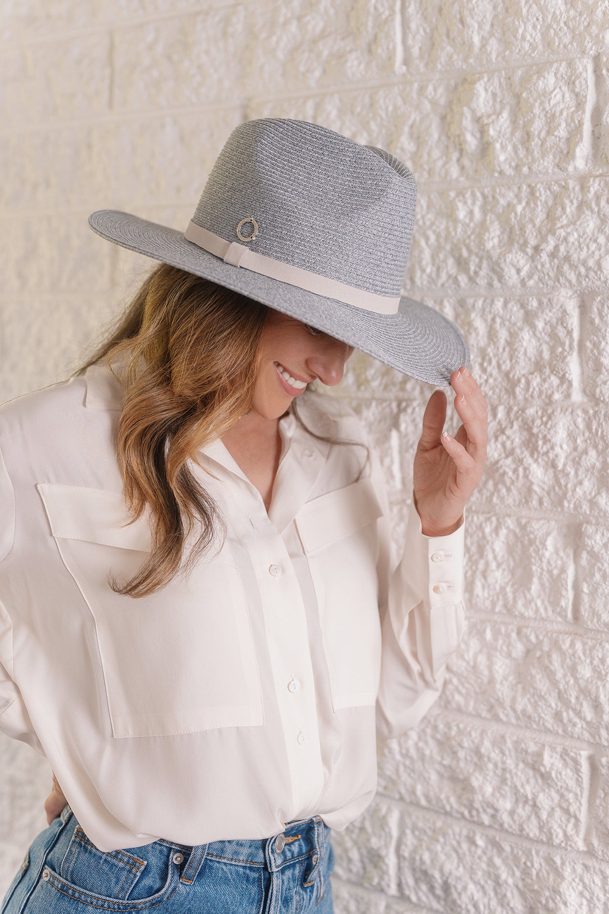 A woman wearing a light gray woven fedora hat with a cream band and a cream button-up shirt, posing against a white textured brick wall.