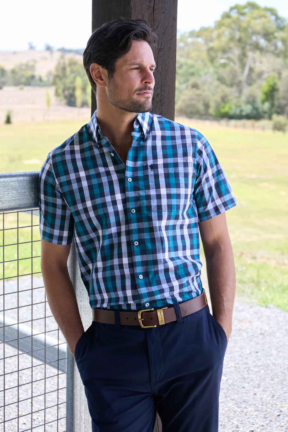 Thomas Cook Men's John S/S Shirt. Man wearing a blue and green checkered shirt with navy pants standing outdoors.