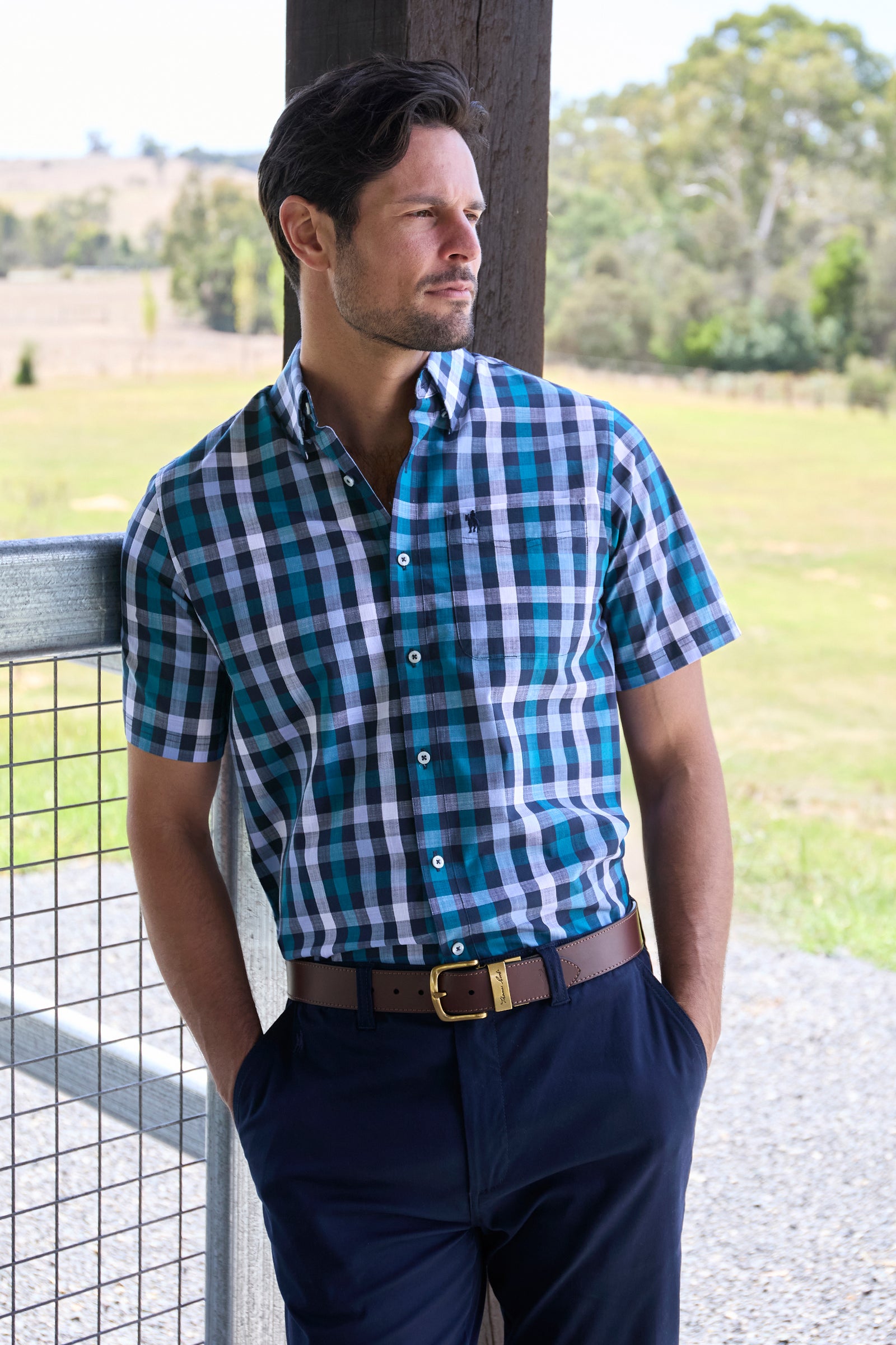 Thomas Cook Men's John S/S Shirt. Man wearing a blue and green checkered shirt with navy pants standing outdoors.