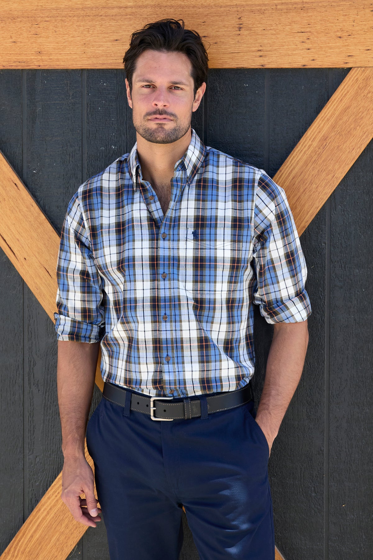 Thomas Cook Men's Daniel L/S Shirt. Man wearing a blue plaid shirt and navy pants standing against a wooden and black background