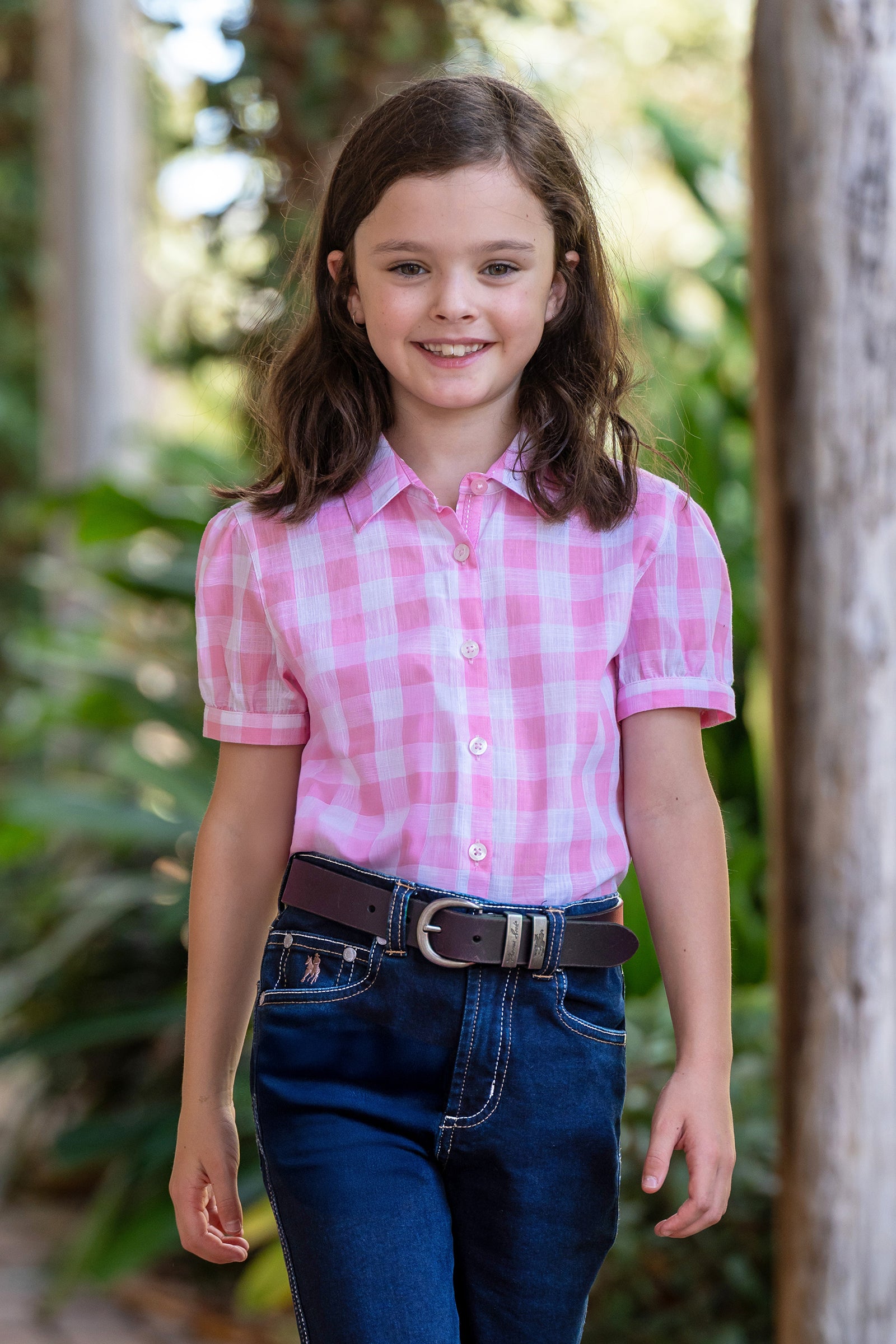Thomas Cook Girl's Eliza S/S Shirt. Young girl wearing a pink checkered shirt and blue jeans standing outdoors with greenery in the background