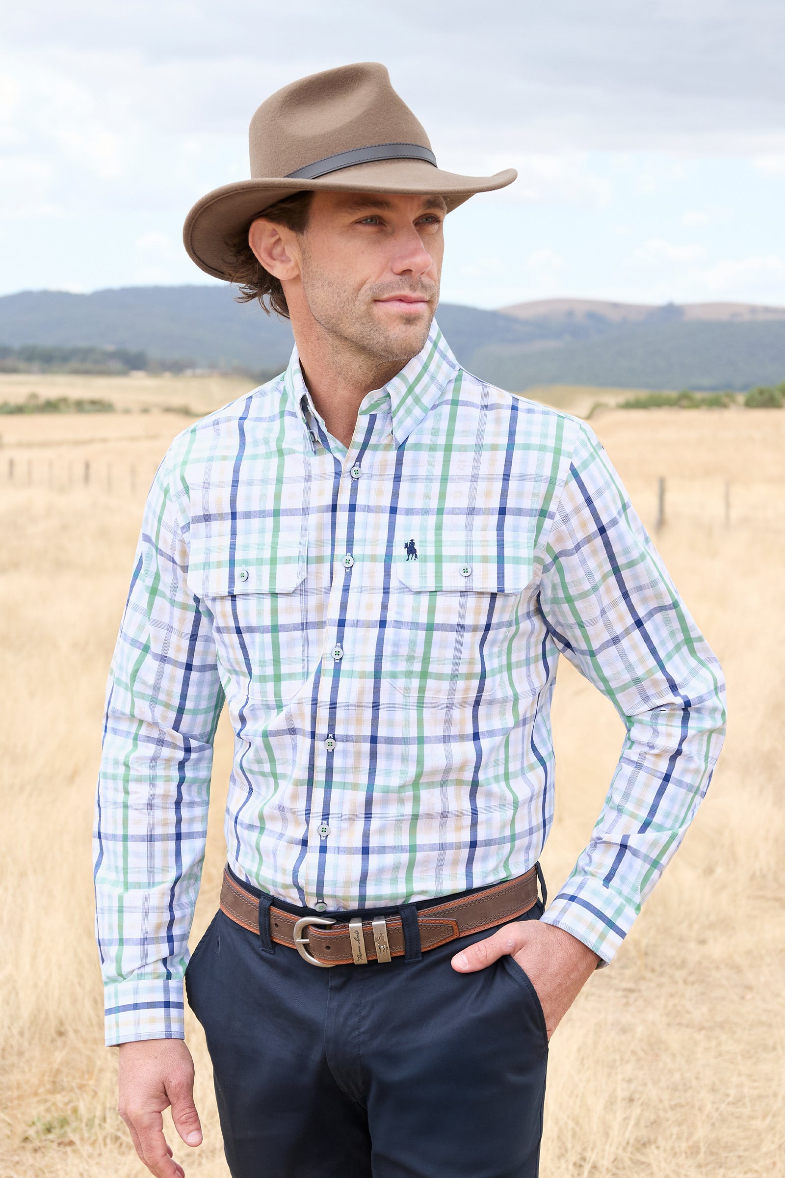 A man wearing a long-sleeve western plaid button-down shirt in white, green, and blue, paired with a brown cowboy hat, dark trousers, and a brown leather belt, standing in a dry grass field.