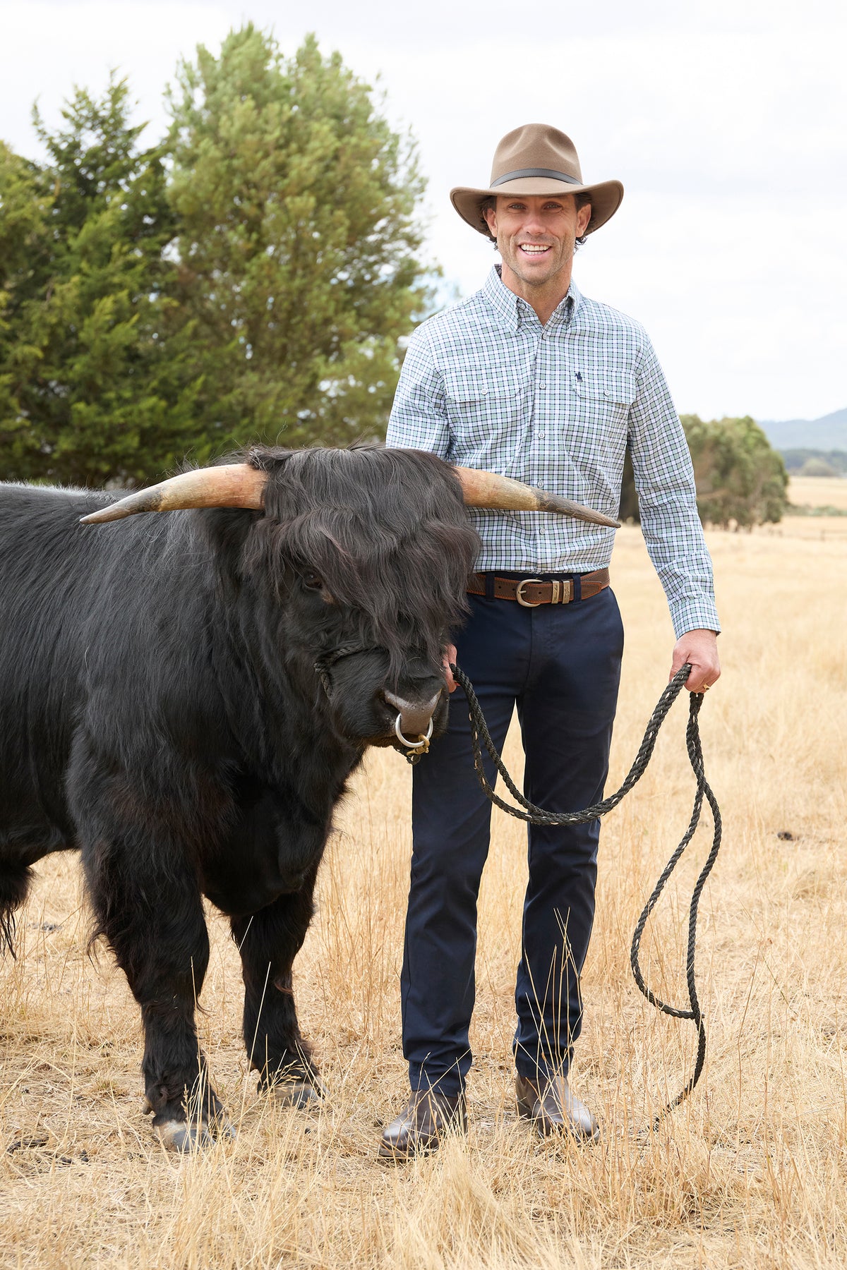 A smiling man in a cowboy hat, light blue plaid shirt, and navy pants stands in a dry grass field holding the reins of a large black long-horn cattle animal, with trees in the background.