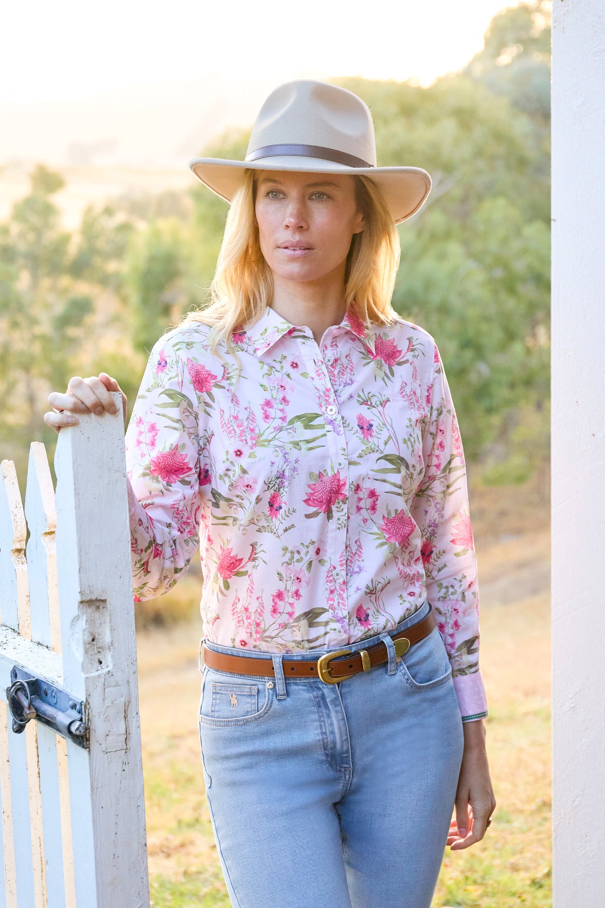 A woman wearing a light pink long-sleeve button-up shirt with a pink and green floral print, styled with light blue jeans, a brown belt, and a beige wide-brim hat, standing outdoors next to a white picket fence.