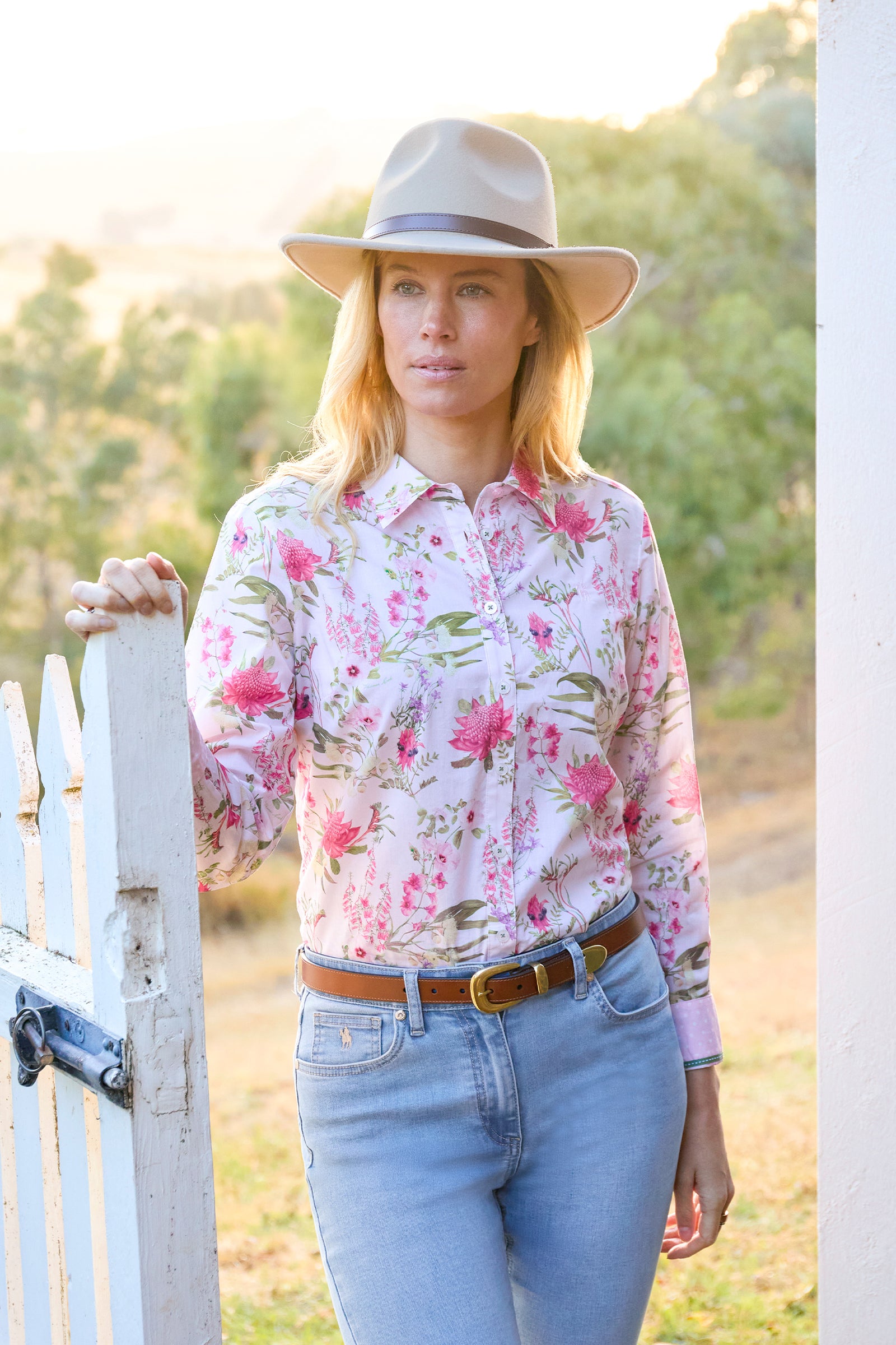 A woman wearing a light pink long-sleeve button-up shirt with a pink and green floral print, styled with light blue jeans, a brown belt, and a beige wide-brim hat, standing outdoors next to a white picket fence.
