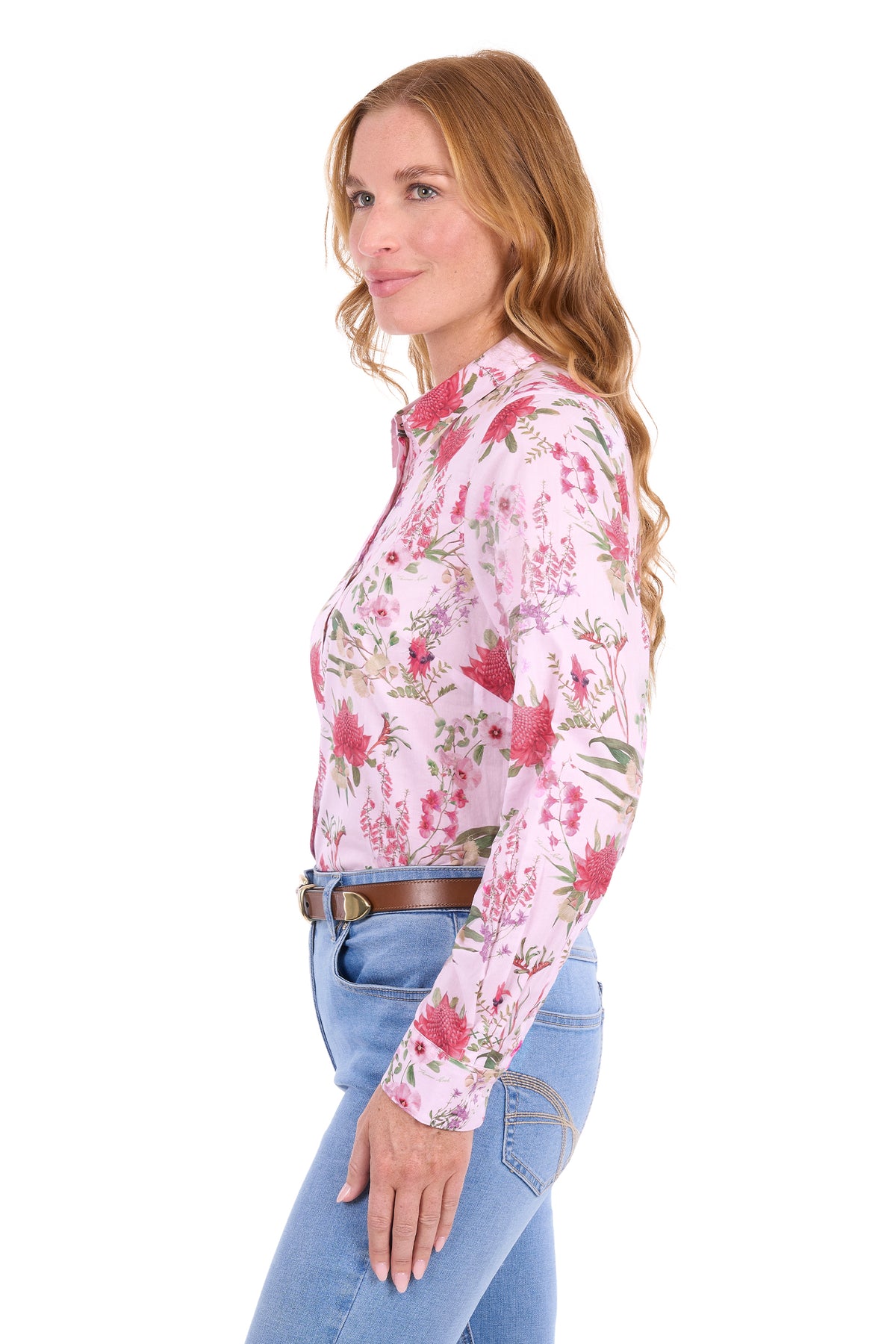 A woman shown in side profile wearing a light pink long-sleeve western-style button-up shirt with a red and green floral print, paired with blue jeans and a brown belt, against a plain white background.