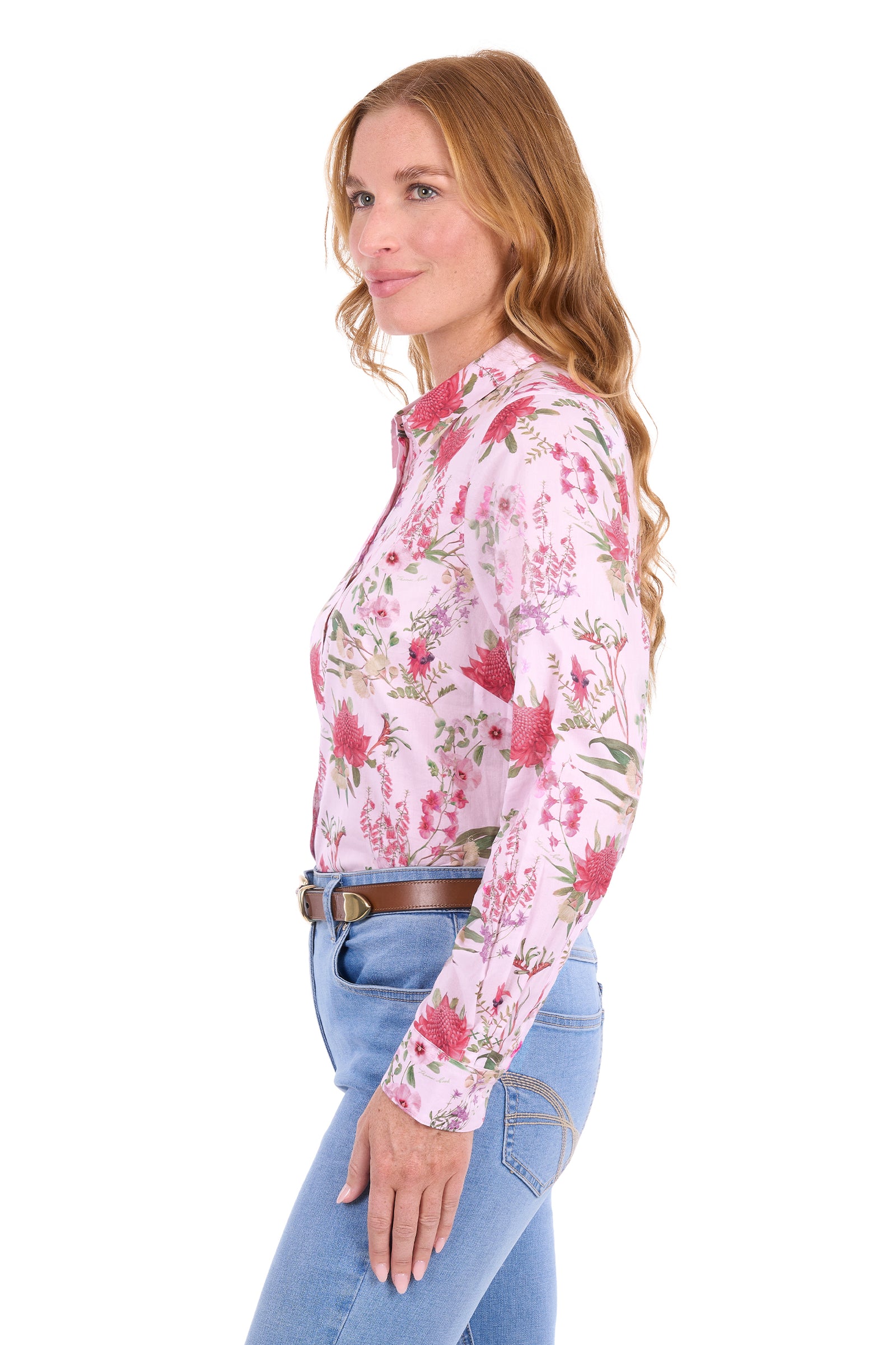 A woman shown in side profile wearing a light pink long-sleeve western-style button-up shirt with a red and green floral print, paired with blue jeans and a brown belt, against a plain white background.