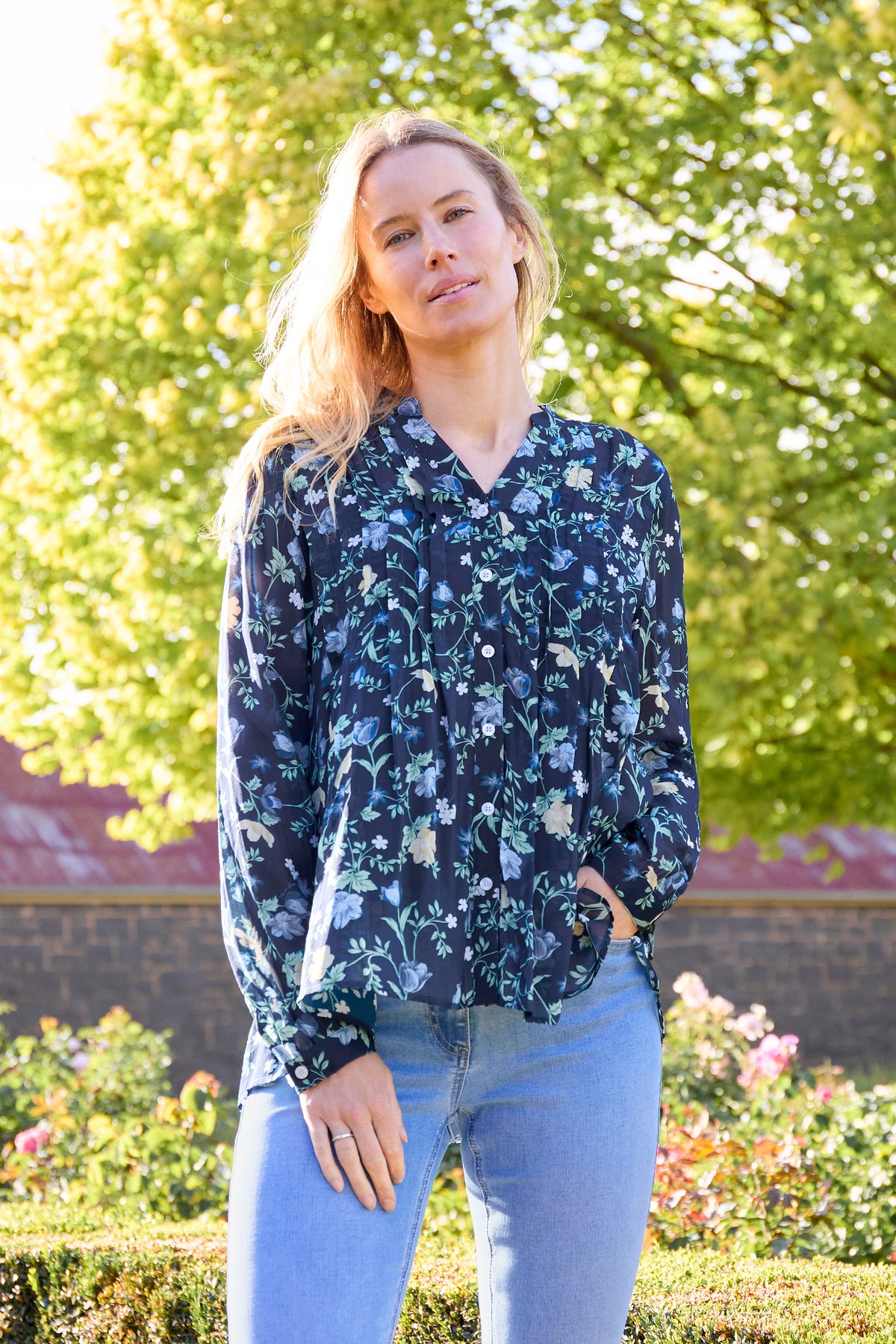 A woman wearing a navy blue long-sleeve blouse with a light blue and white floral pattern, paired with light blue jeans, standing outdoors in front of a green garden background.