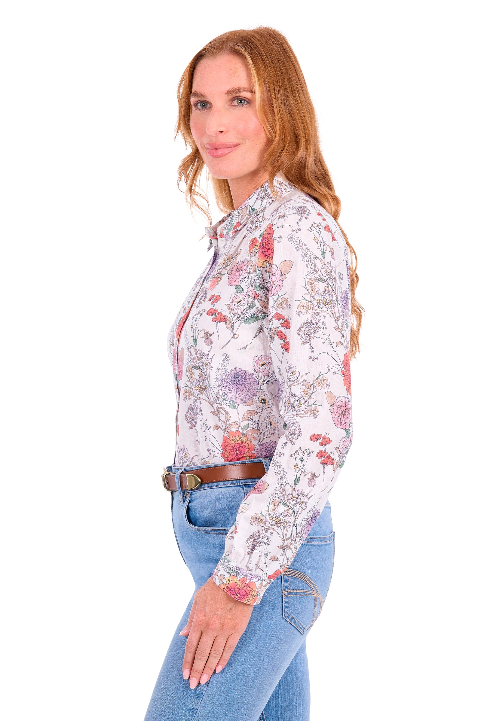 A woman modeling a long-sleeve western-style button-up shirt with a multicolor floral print on a white base, paired with blue jeans and a brown belt, against a plain white background.