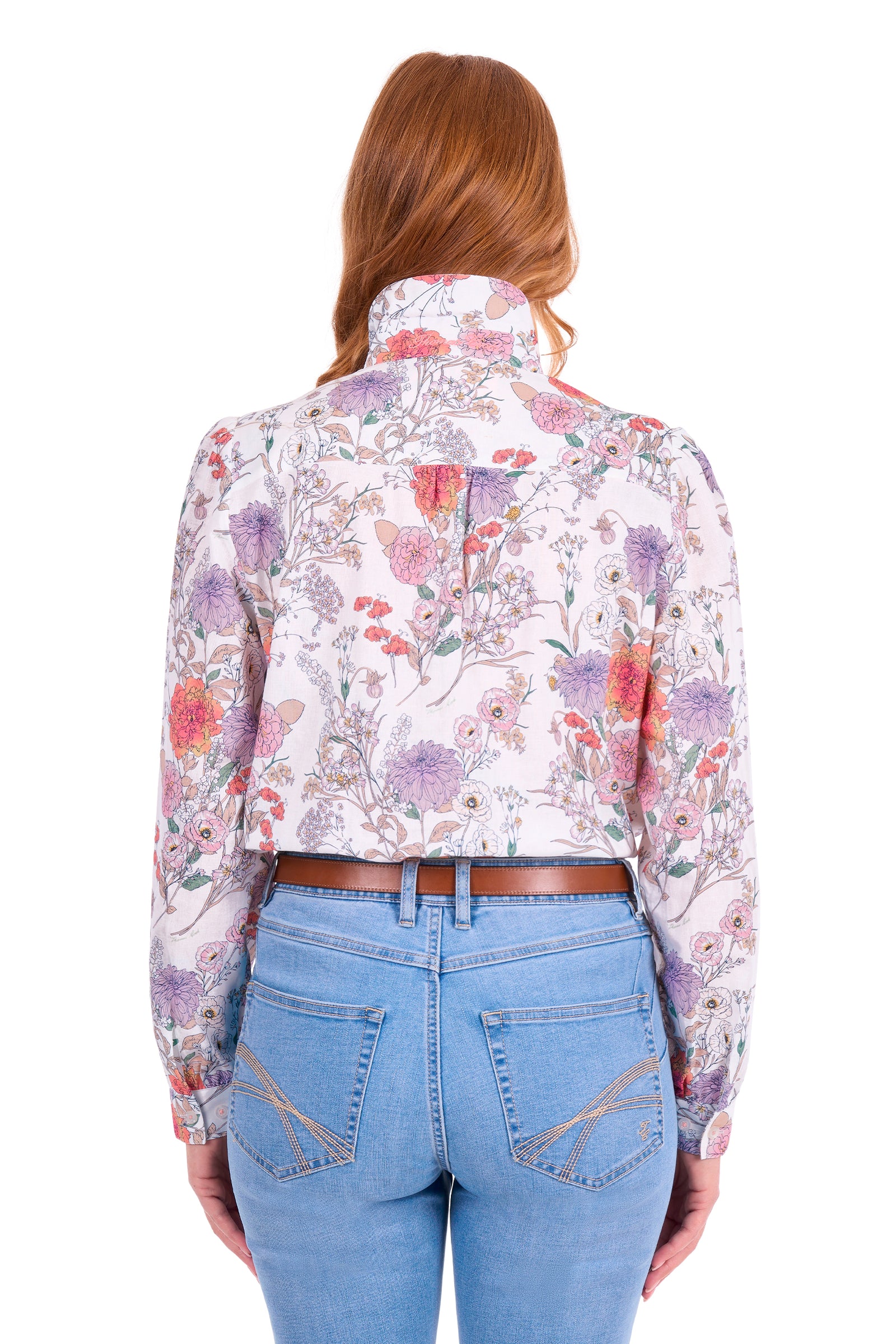 A woman viewed from the back wearing a long-sleeve collared blouse with a dense floral print in pink, purple, and green on a light background, paired with light blue jeans and a brown belt, against a plain white studio background.