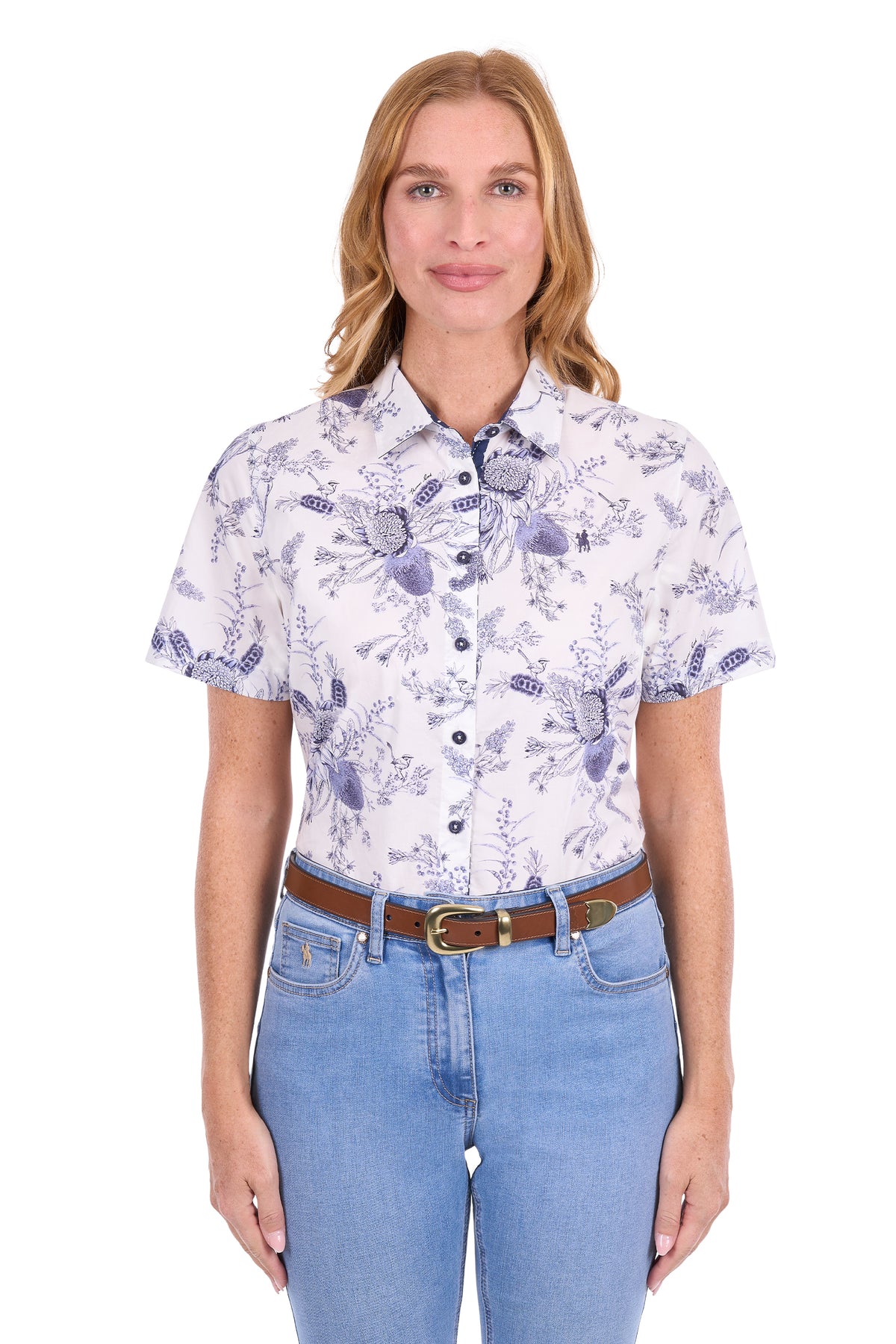 A woman modeling a short-sleeve button-up shirt with a white base and a blue floral and butterfly pattern, paired with light blue jeans and a brown belt, against a plain white background.