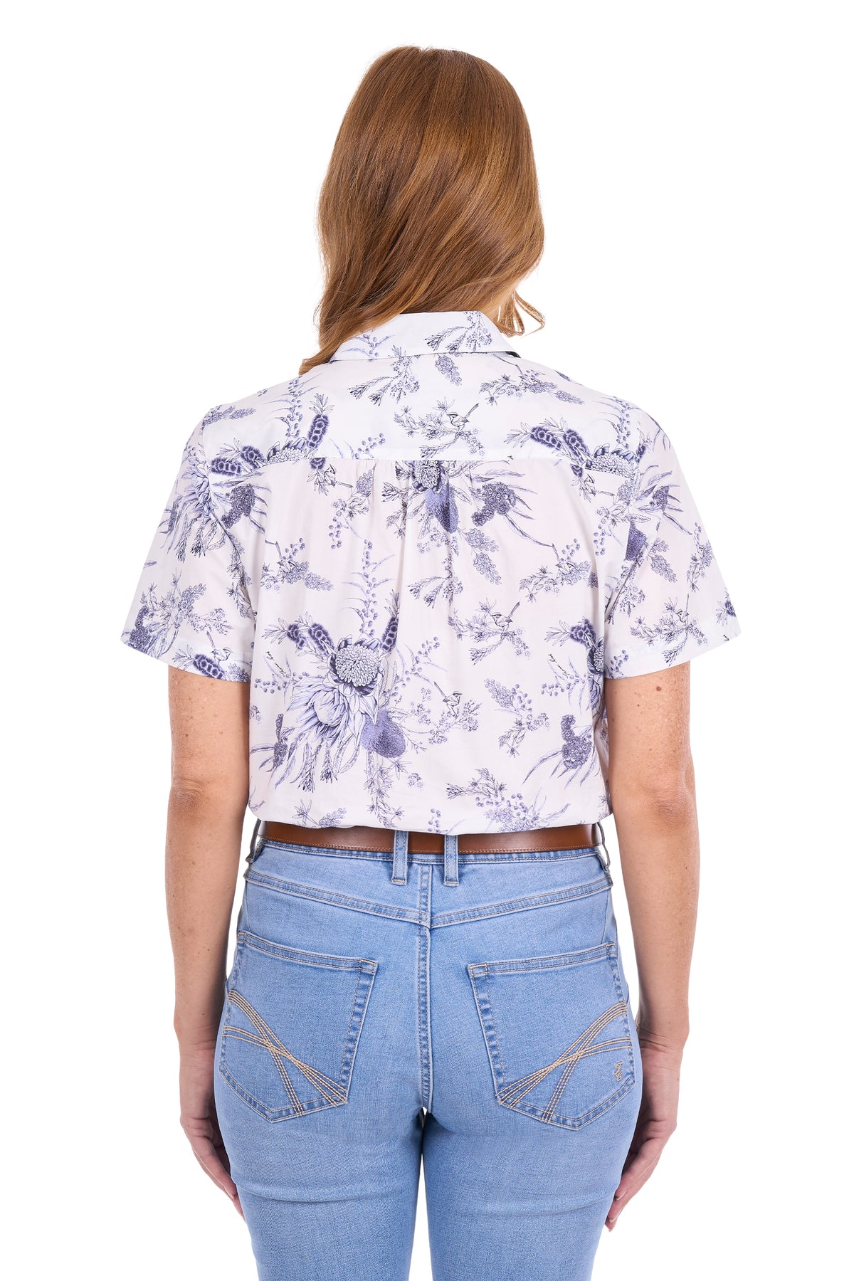 A woman viewed from the back wearing a short-sleeve white button-up shirt with a purple floral print and a bow at the back collar, paired with light blue jeans against a white studio background.