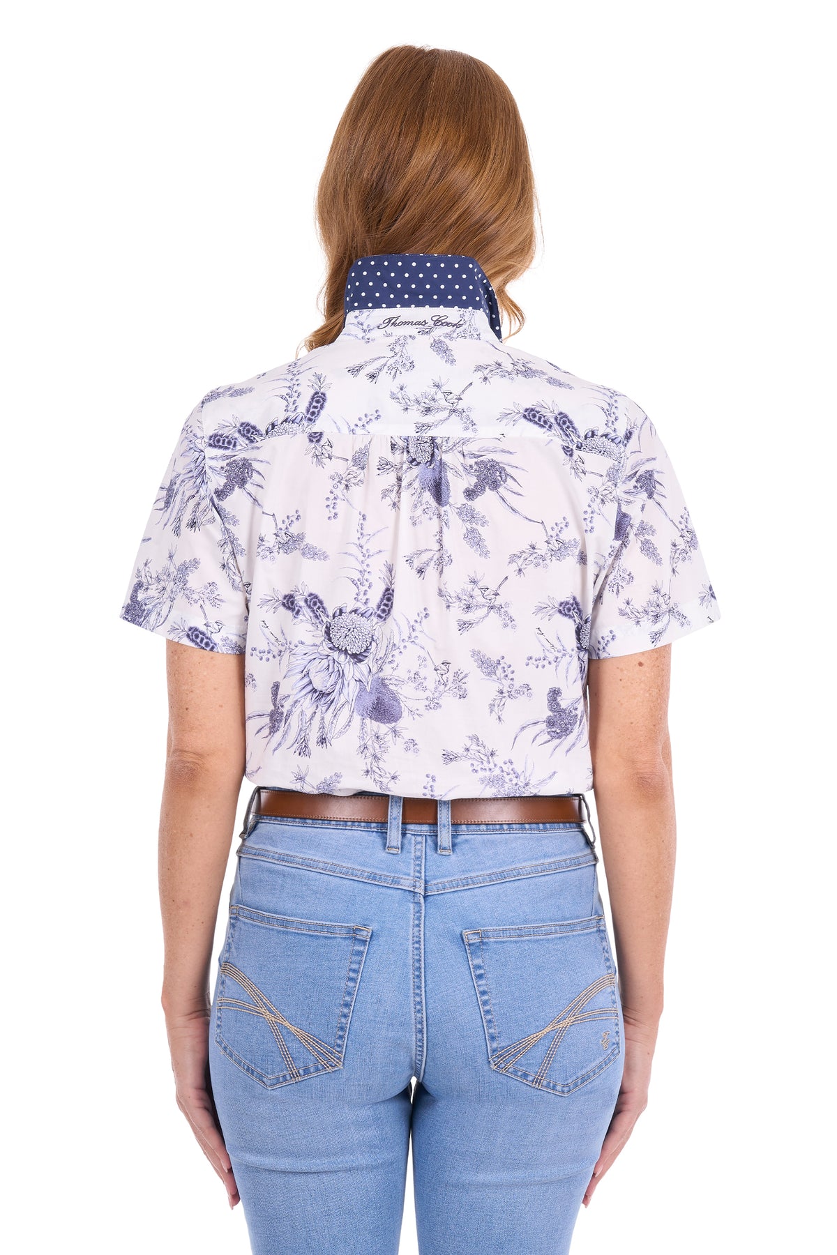 A woman viewed from the back wearing a short-sleeve collared button-up shirt with a blue and white floral toile pattern, paired with light blue jeans and a brown belt, against a plain white background.