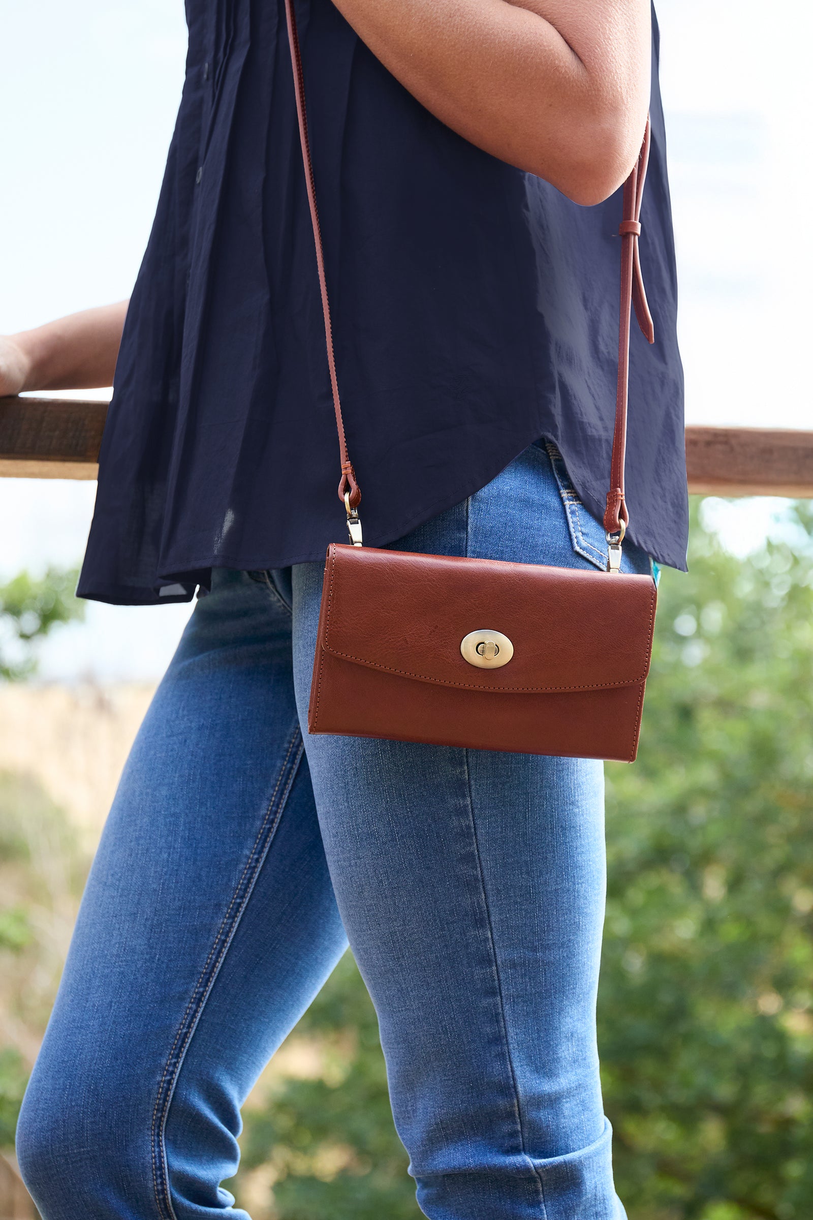 A brown leather crossbody handbag with a gold-tone clasp, worn by a person wearing a navy blue top and blue jeans.