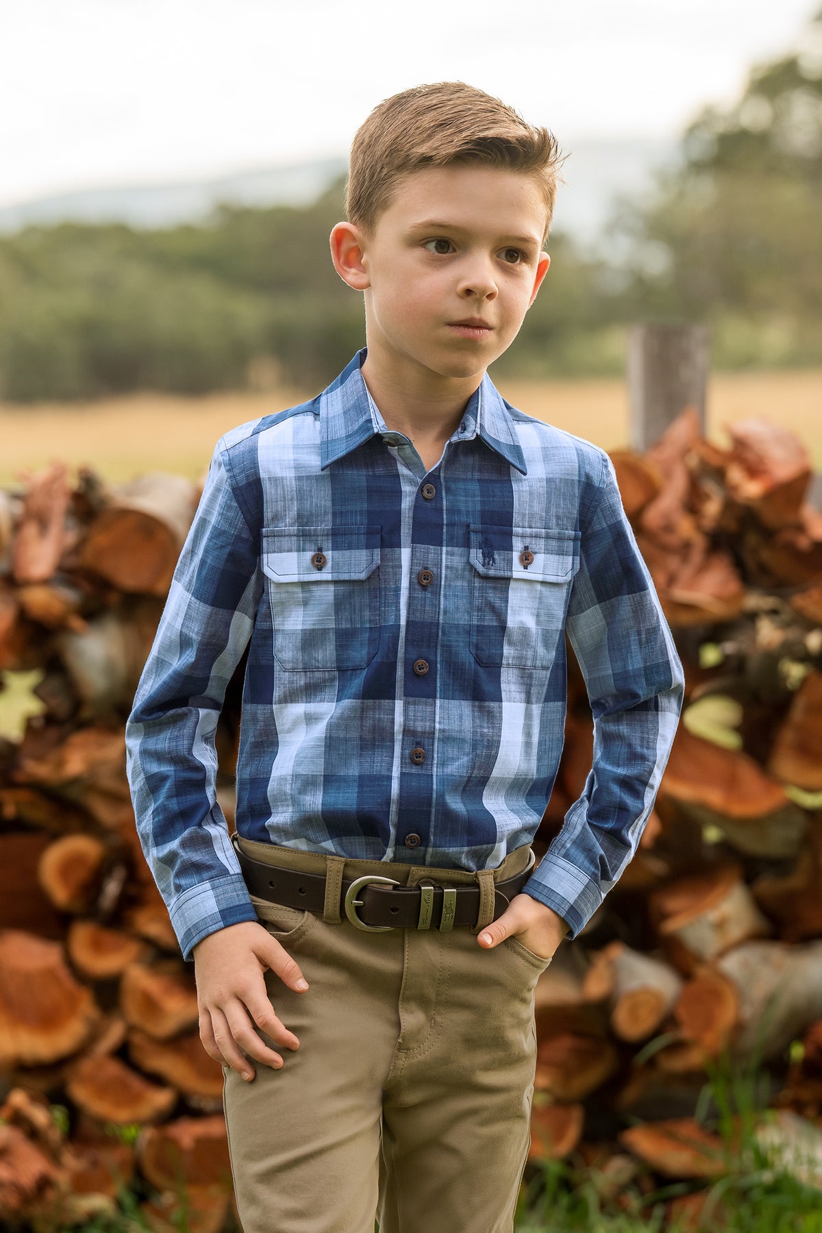 A young boy wearing a blue and navy plaid long-sleeve button-up shirt with two chest pockets, paired with khaki pants and a brown belt, standing outdoors in front of a stack of cut logs.