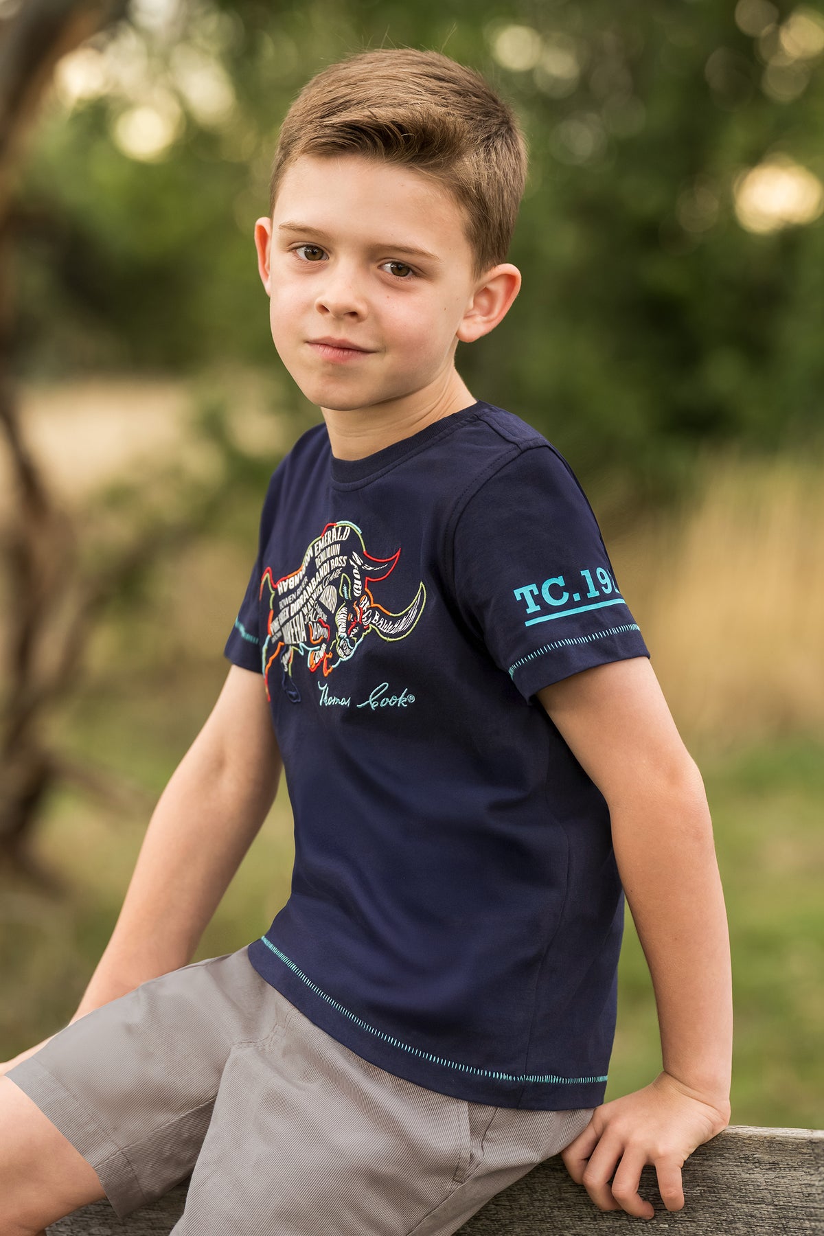 A young boy wearing a navy blue short-sleeve t-shirt with a graphic print on the front and 'TC.19' on the sleeve, sitting outdoors on a stone ledge.