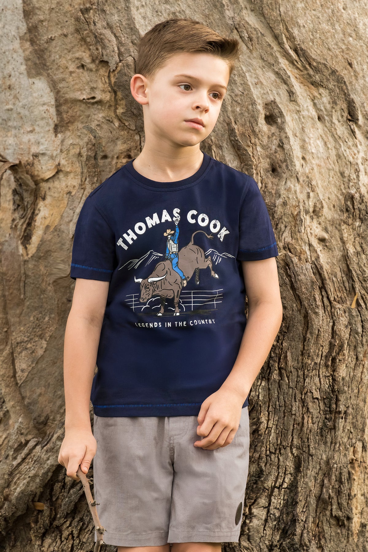 A young boy wearing a navy blue graphic t-shirt with 'THOMAS COOK' printed on the front, paired with gray shorts, standing in front of a large textured tree trunk.