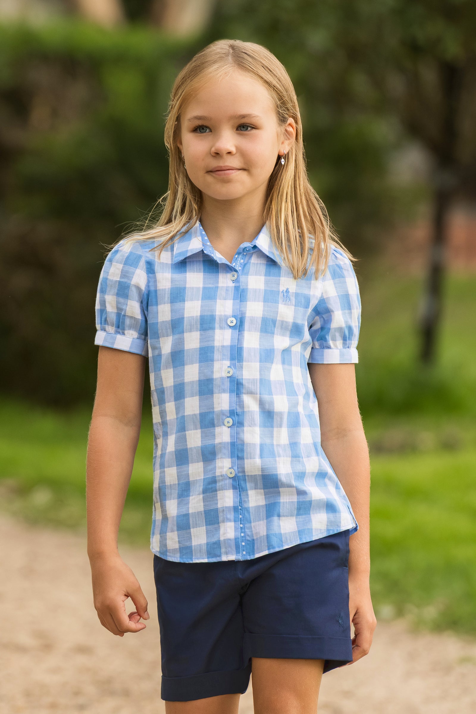 A young girl with blonde hair stands outdoors wearing a light blue and white gingham short-sleeve button-up shirt and navy blue shorts.