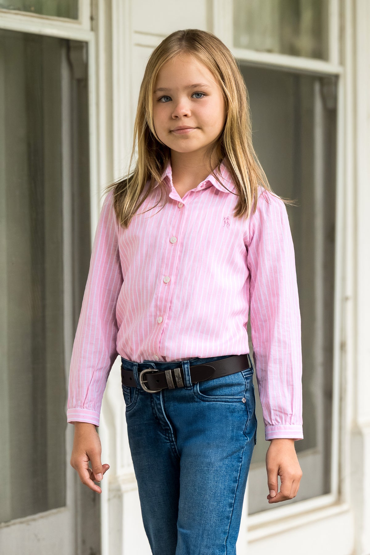 A young girl with blonde hair stands in front of a white paneled door, wearing a pink and white vertically striped long-sleeve button-down shirt, blue jeans, and a black belt.