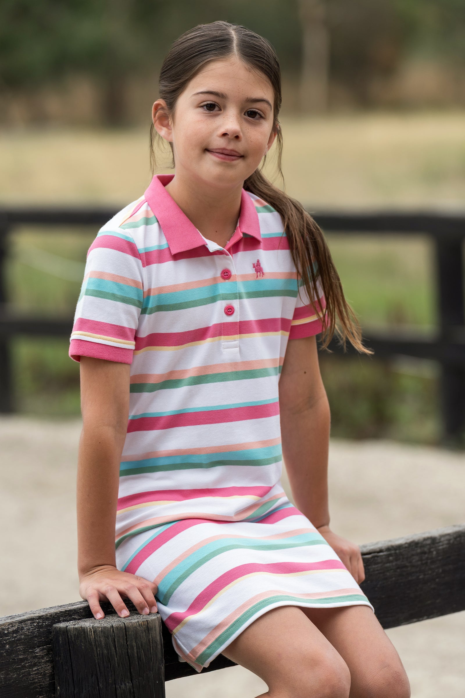 A young girl with long brown hair sitting on a wooden fence, wearing a short-sleeve polo dress with horizontal pink, teal, and light green stripes on a white background.