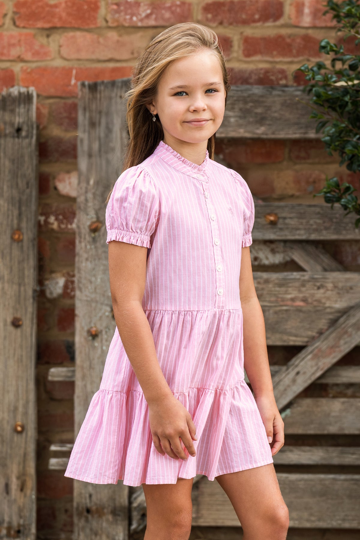 A young girl with blonde hair wearing a pink and white vertically striped tiered dress, standing in front of a rustic wooden door and brick wall.