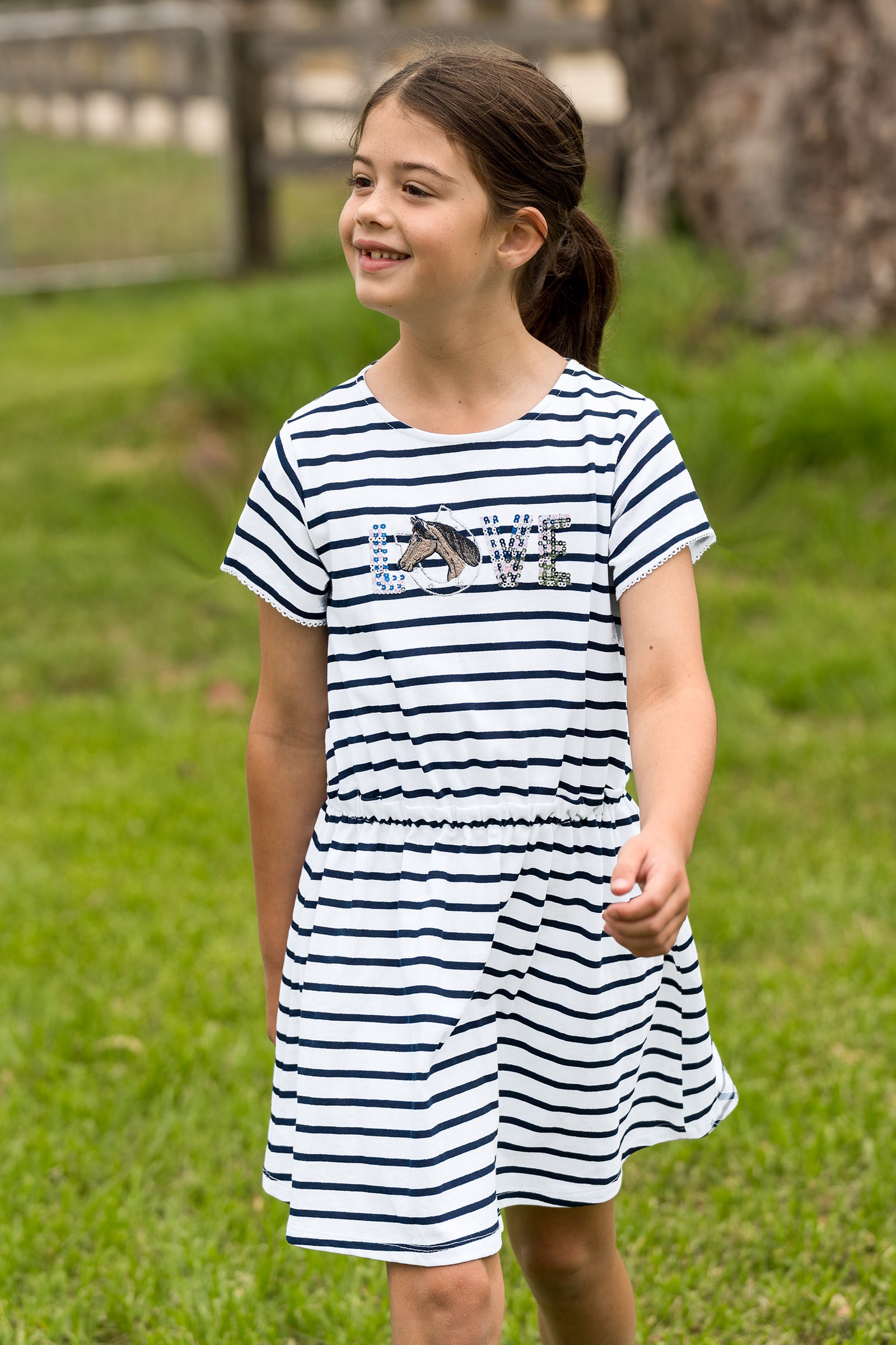 A young girl wearing a navy and white horizontally striped short-sleeve dress with a 'LOVE' graphic across the chest, standing outdoors on a green grassy lawn.