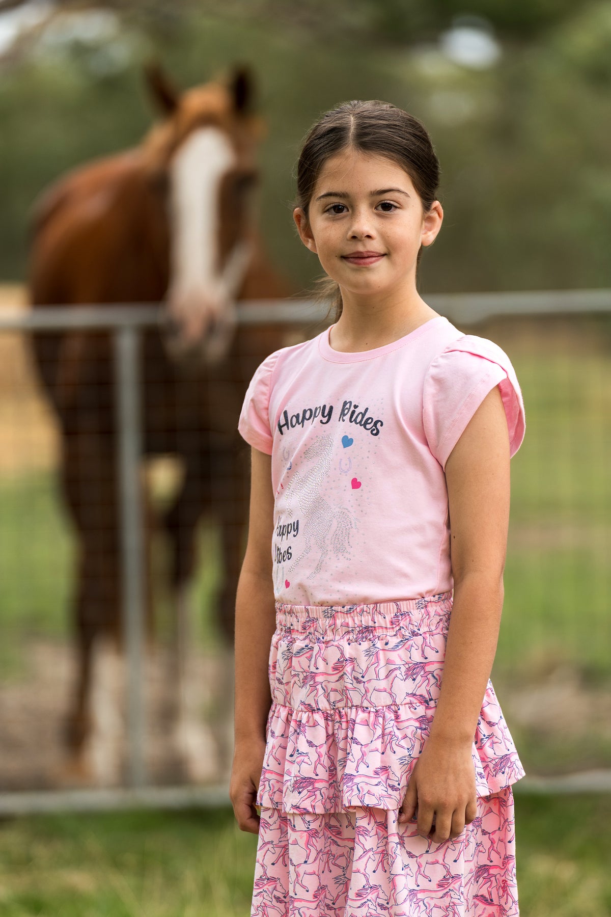 A young girl stands in front of a horse fence wearing a light pink t-shirt with 'Happy Rides' printed on it and a matching pink patterned tiered skirt.
