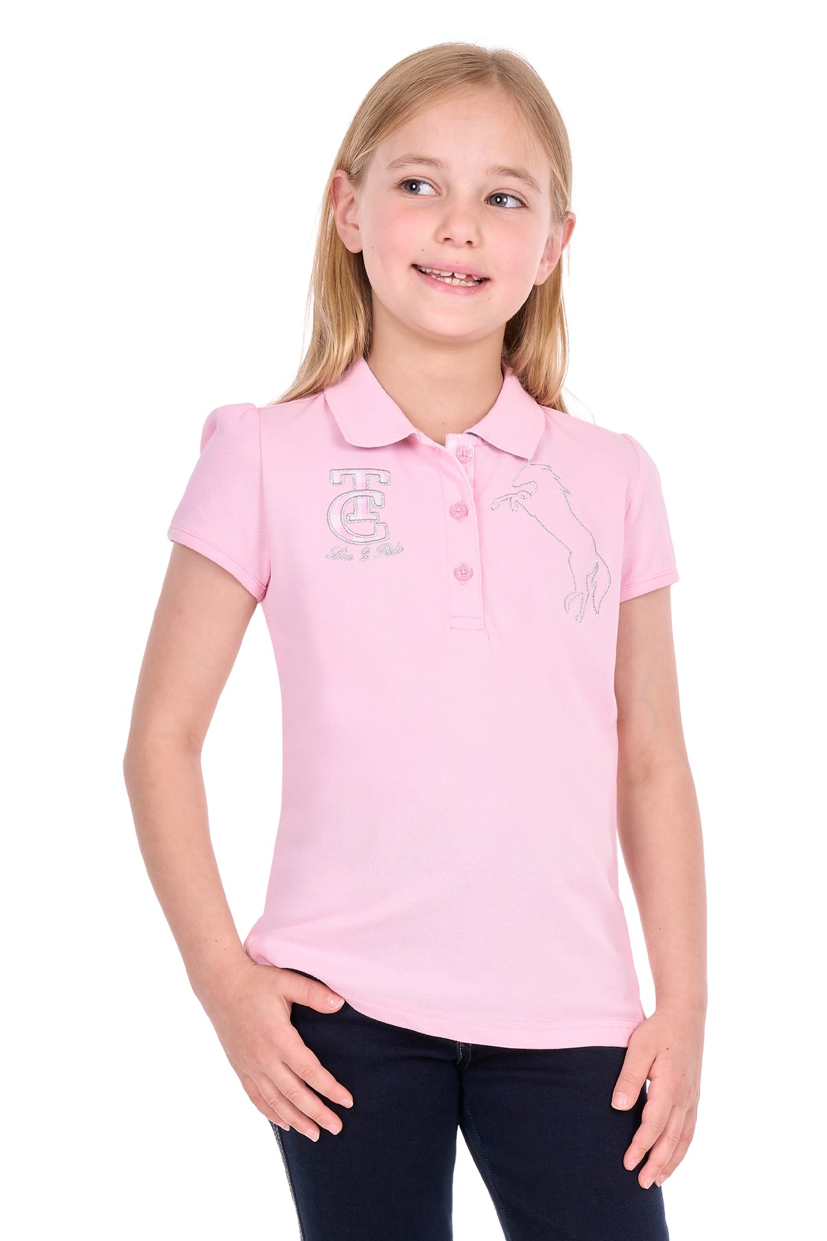 A young girl with blonde hair wearing a light pink polo shirt with a silver-toned embroidered crest on the left chest and dark navy pants, standing against a plain white background.