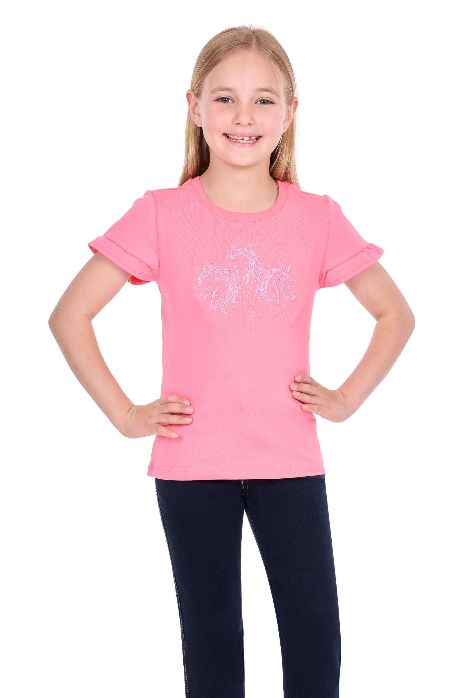 A young girl with blonde hair smiling and posing with her hands on her hips, wearing a bright pink short-sleeve t-shirt with a subtle white graphic on the front and dark navy pants, set against a plain white background.