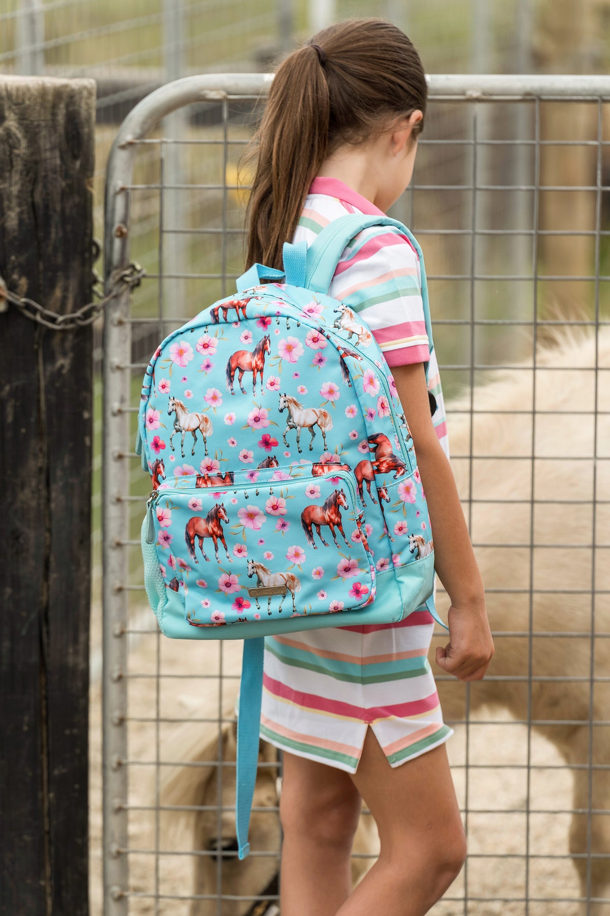 A child wearing a light blue backpack with a horse and flower print, standing in front of a metal fence at an outdoor ranch.