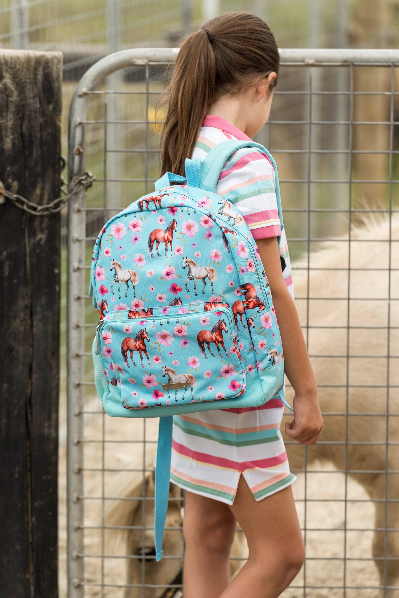 A child wearing a light blue backpack with a horse and flower print, standing in front of a metal fence at an outdoor ranch.