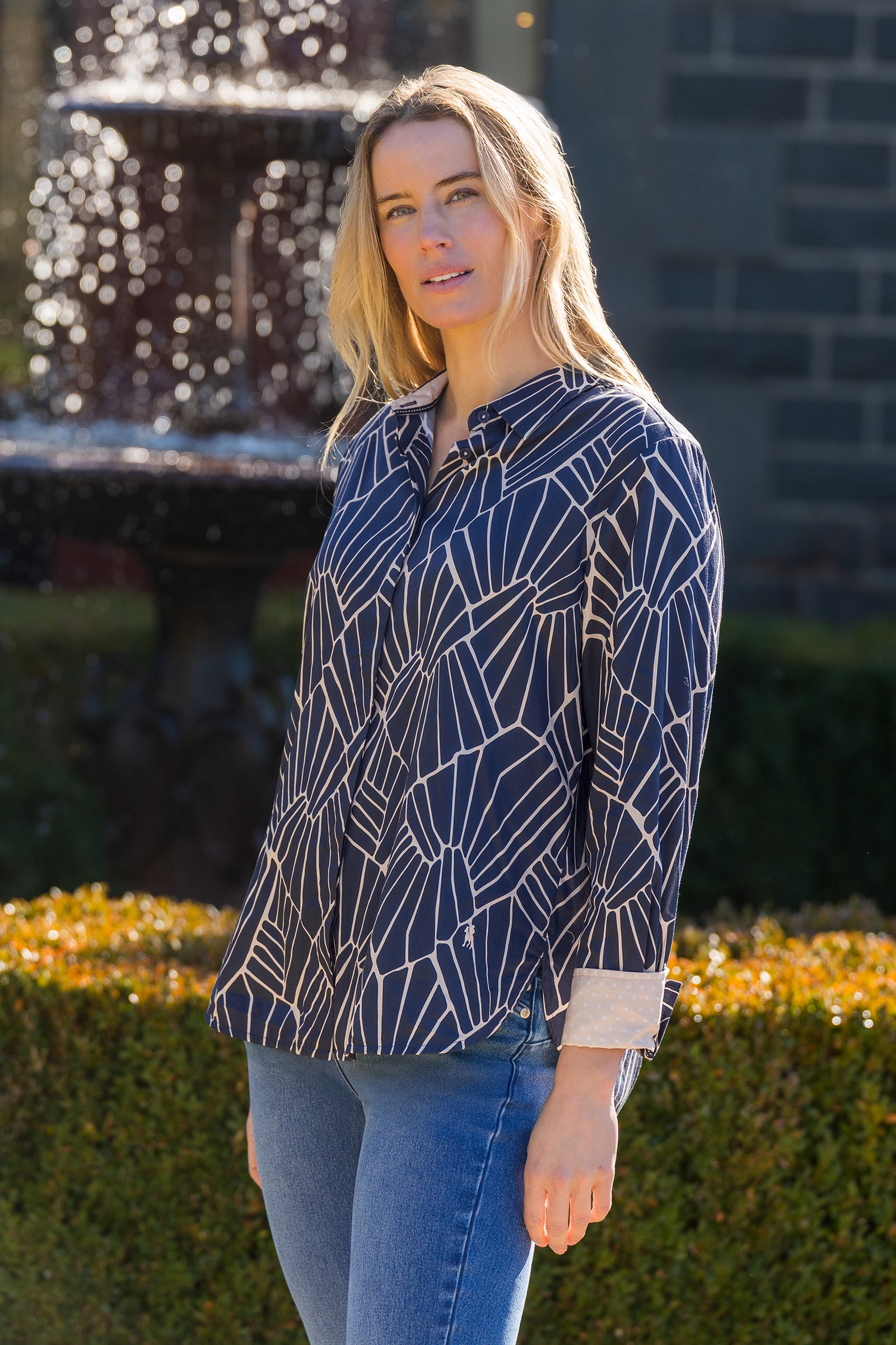 Woman wearing a navy blue patterned shirt and jeans standing in front of a fountain.