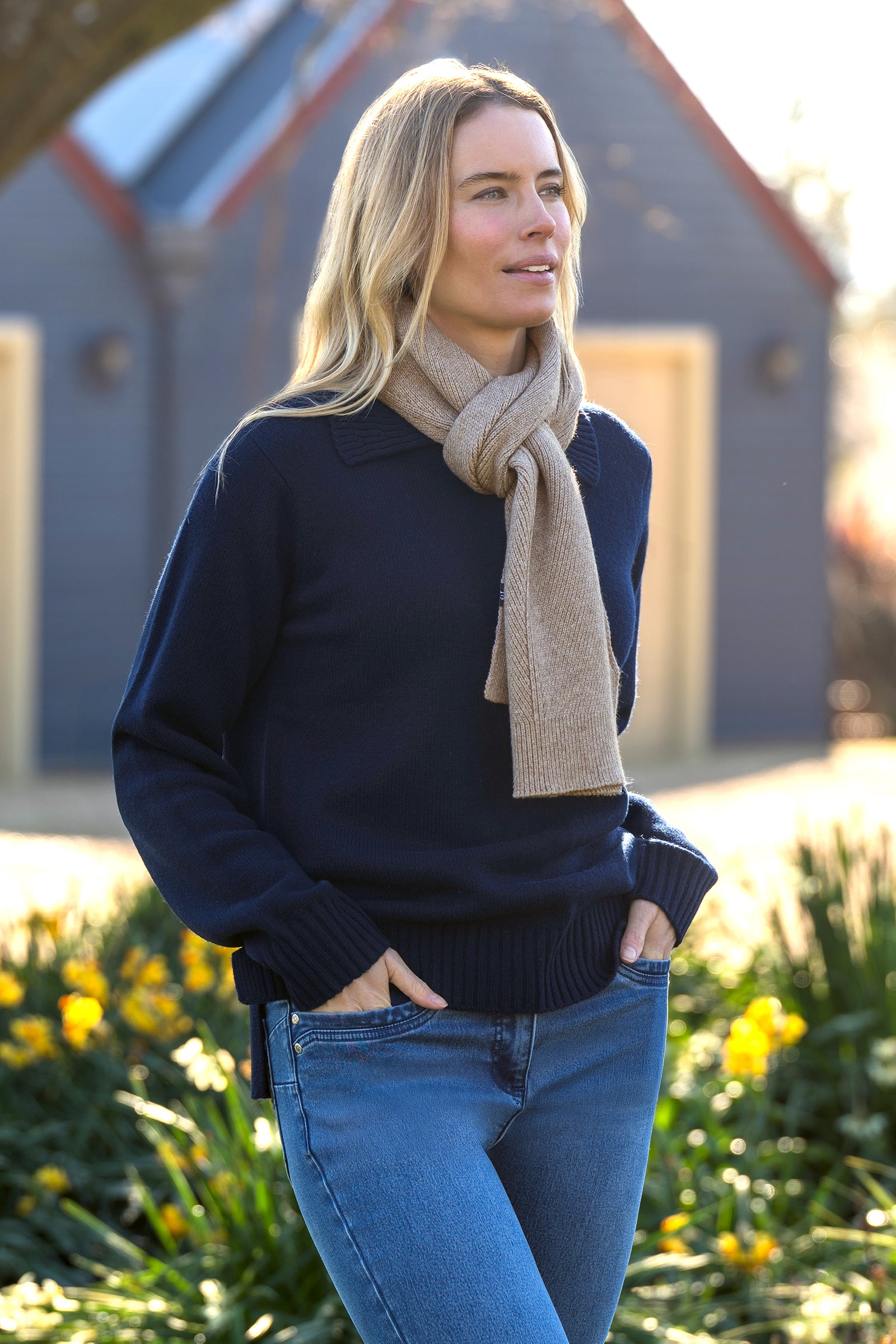 Woman wearing a navy sweater and beige scarf standing outdoors with flowers and a building in the background