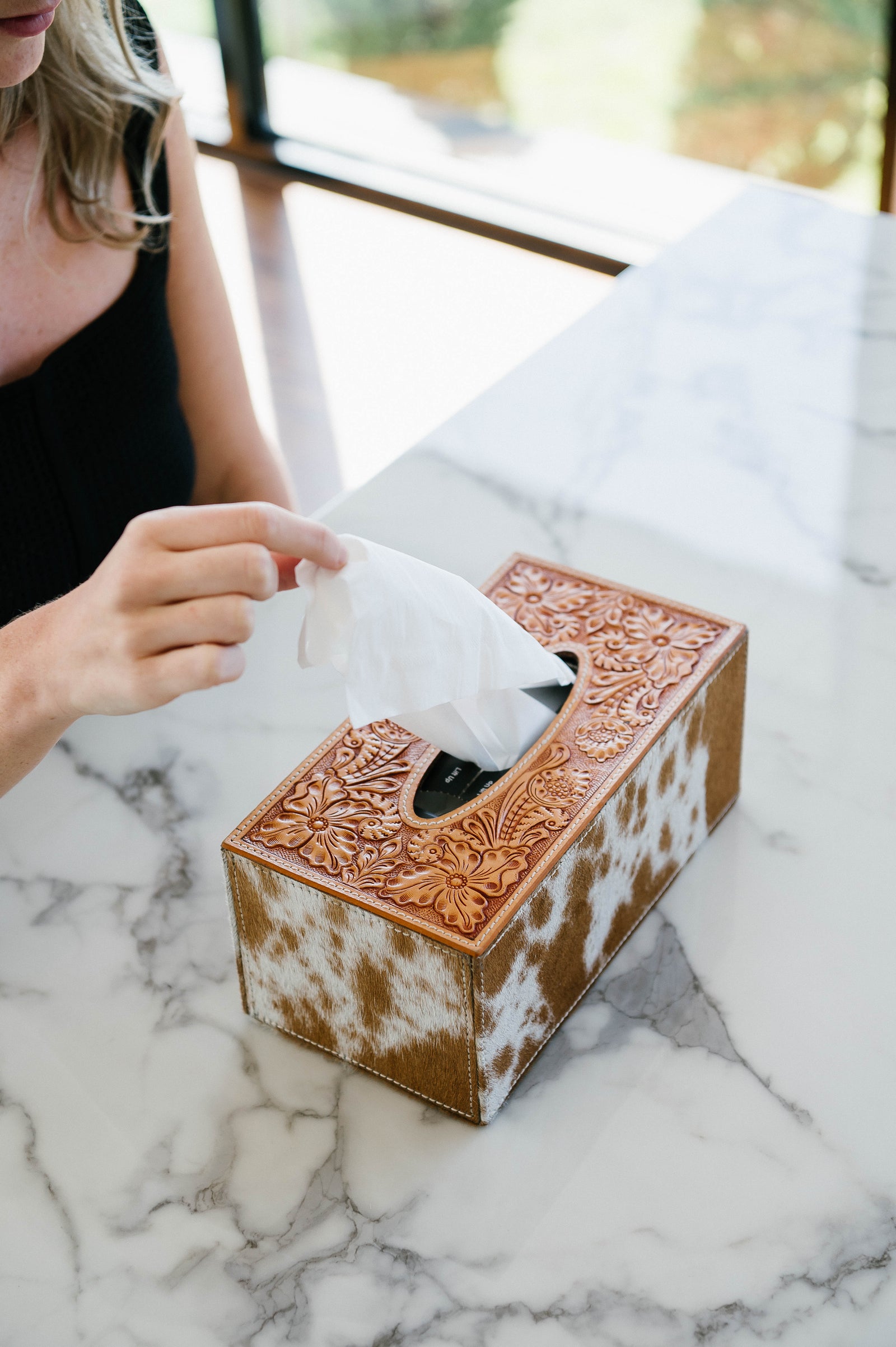 Person taking a tissue from a decorative cowhide-patterned tissue box on a marble surface.