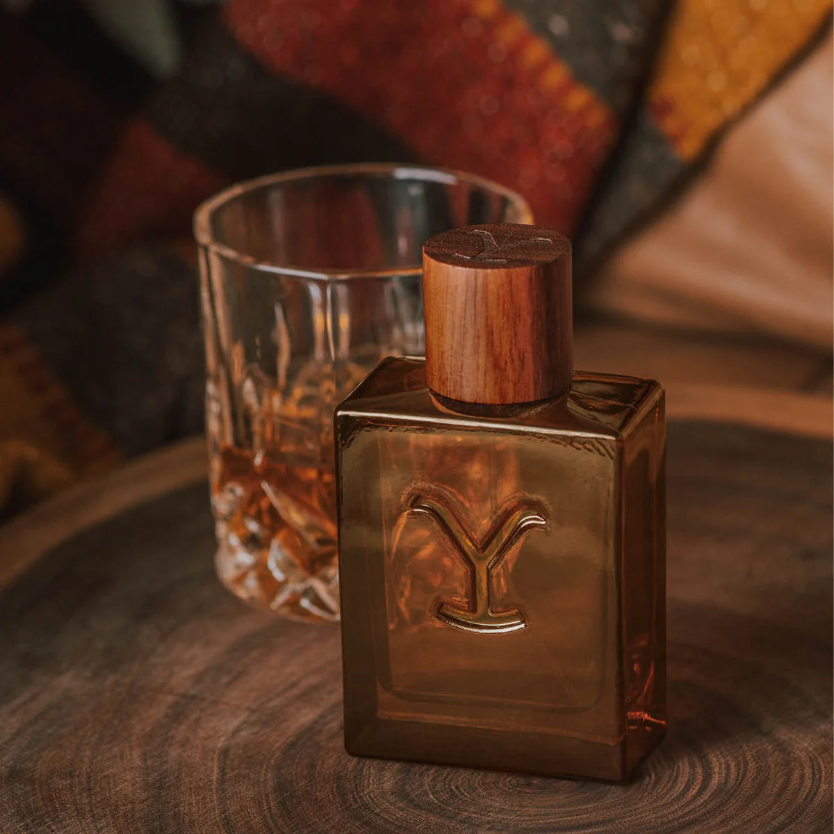 A square amber-brown glass bottle with a carved wooden cap and an embossed letter Y on the front, placed on a wooden surface beside a cut-glass tumbler.