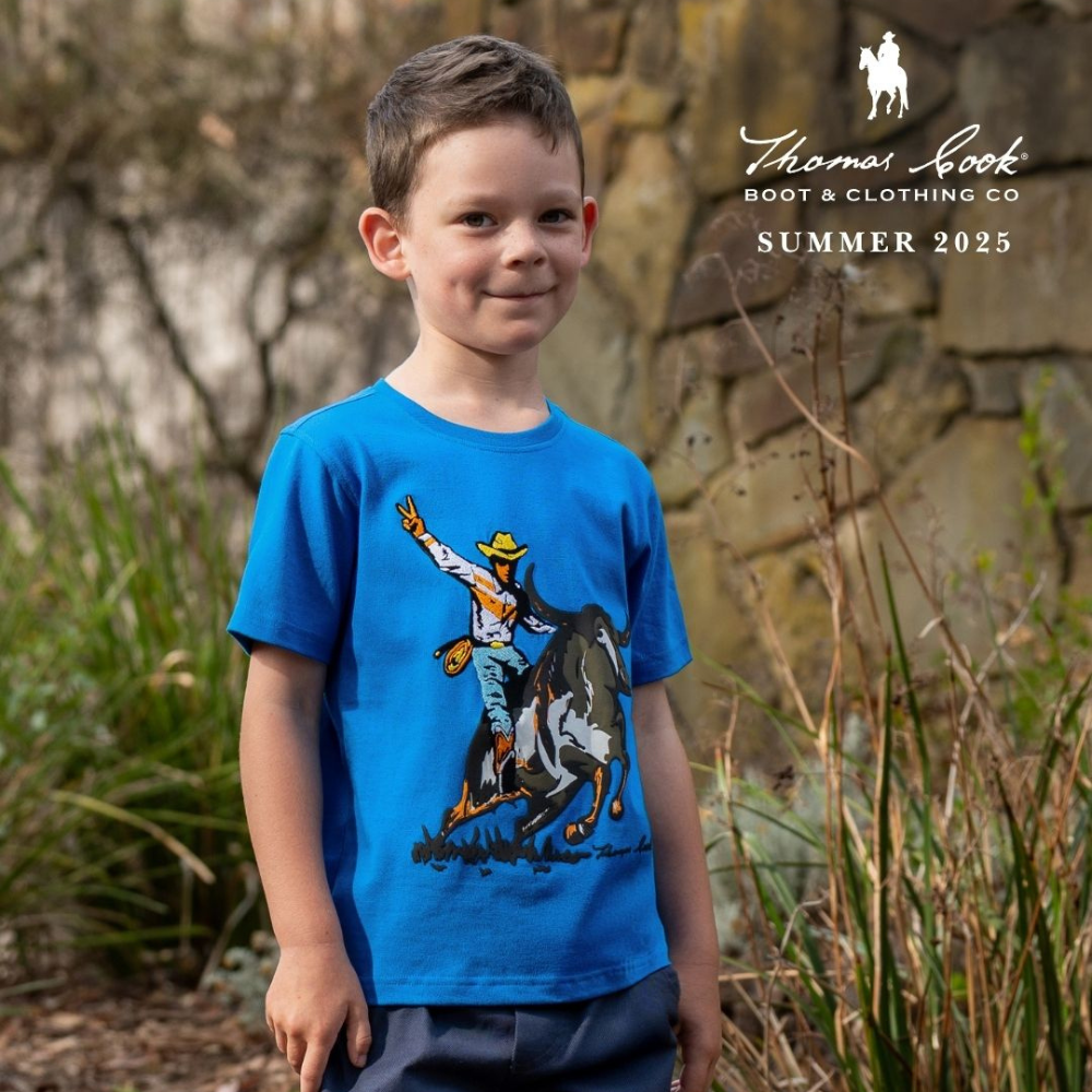 Thomas Cook Boy's Summer. Young boy wearing a blue t-shirt with a graphic design, standing outdoors with a stone wall and plants in the background. Thomas Cook Boot & Clothing Co logo and text visible.
