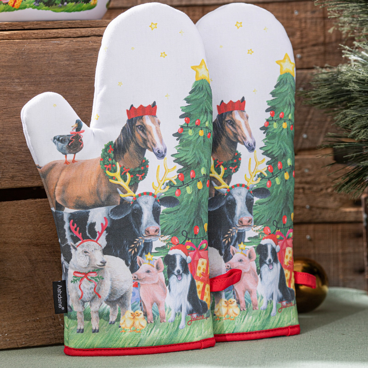 Two white oven mitts with a festive Christmas farm illustration, featuring a horse, cows, sheep, pig, dog, and decorated Christmas trees, set against a rustic wooden background.