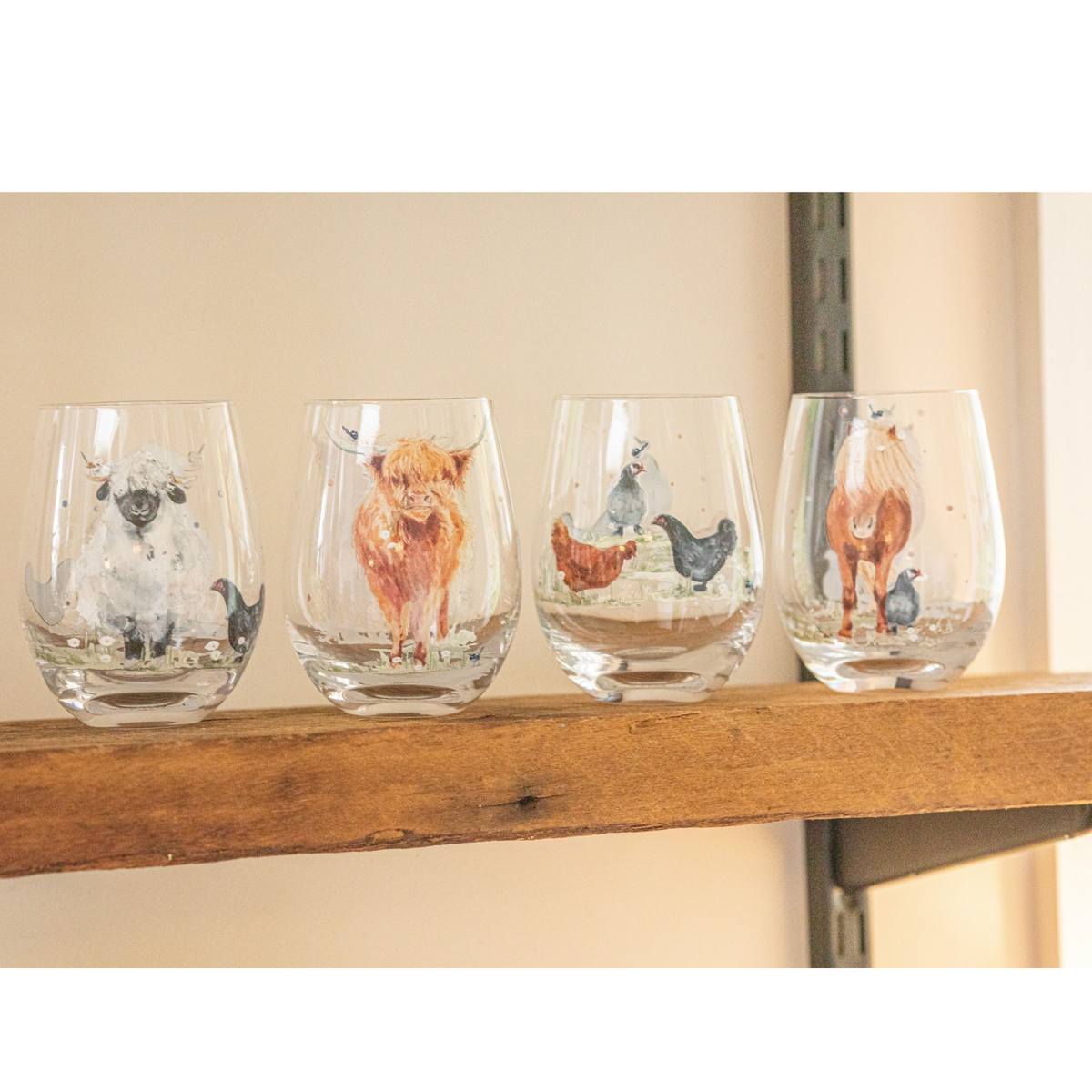 Four clear stemless wine glasses with hand-painted farm animal illustrations, displayed on a rustic wooden shelf against a beige wall.