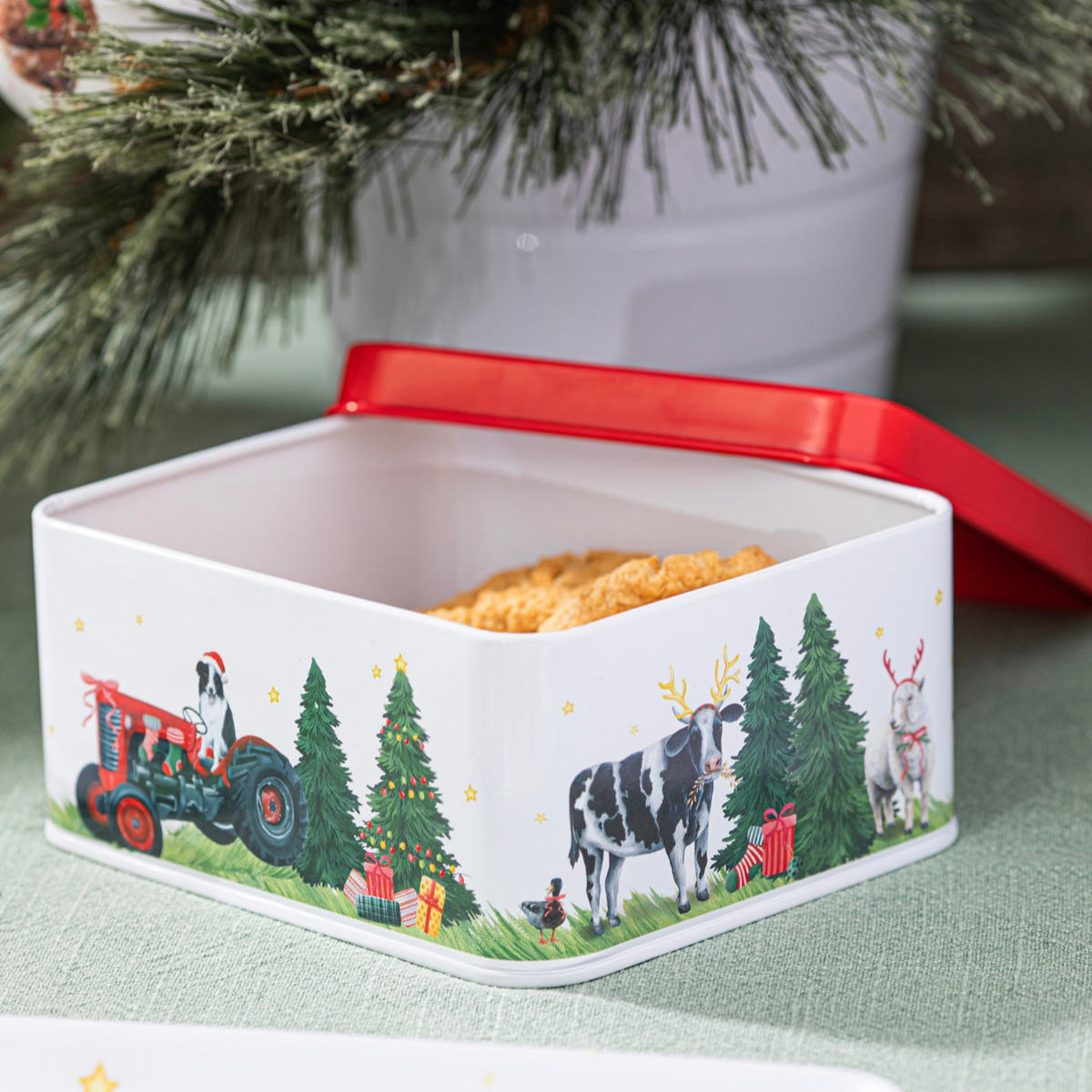 A square white metal tin with a festive Christmas-themed border illustration, shown open with its red lid resting beside it. The tin contains a golden-brown baked good, and the background features a blurred Christmas tree and green tablecloth.