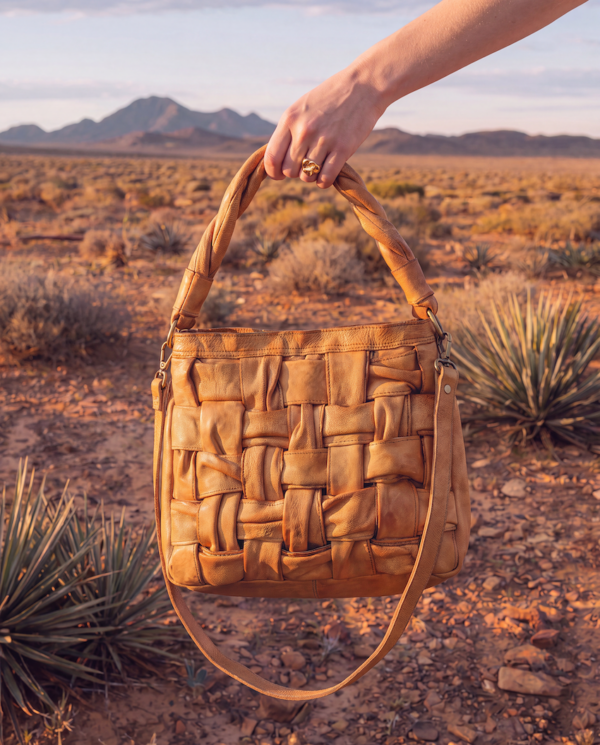 A tan woven leather handbag held by a hand against a desert landscape background with mountains and yucca plants.