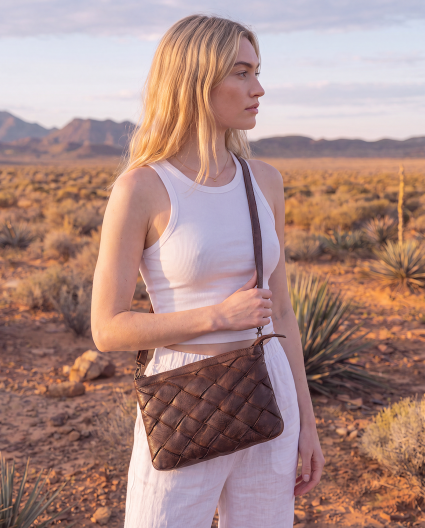 A woman stands in a desert landscape wearing a brown quilted leather crossbody bag with a matching strap. The background features sandy terrain, desert plants, and distant mountains under a warm sunset sky.