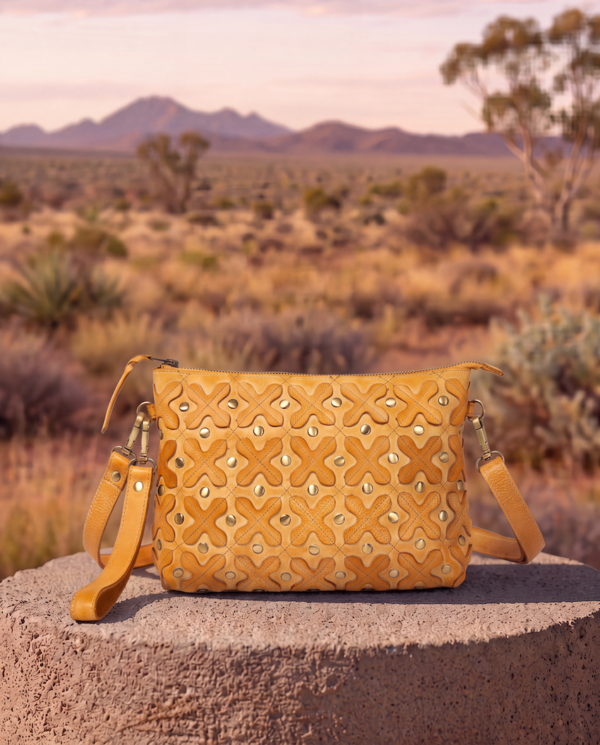 A mustard yellow embossed leather shoulder bag with geometric studs, displayed on a stone pedestal in an outdoor desert landscape at sunset.