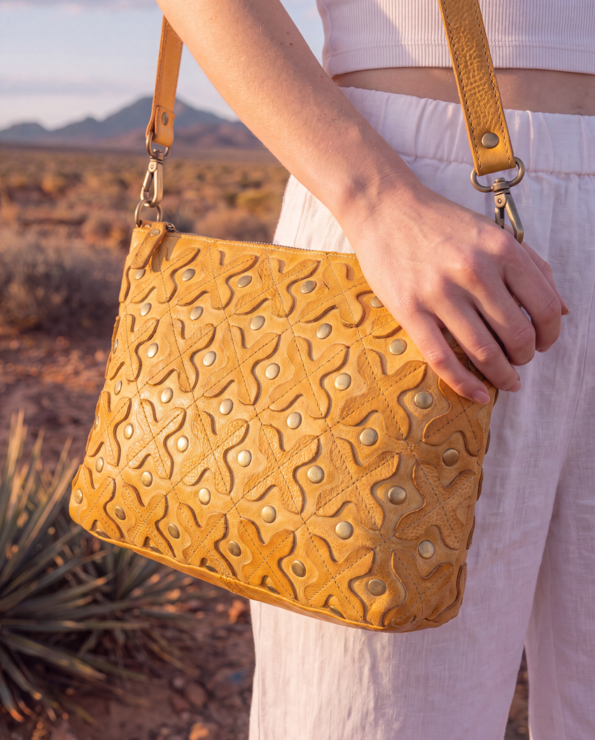 A mustard yellow textured leather shoulder bag with a starburst pattern and brass studs, held by a person wearing white pants in a desert landscape.