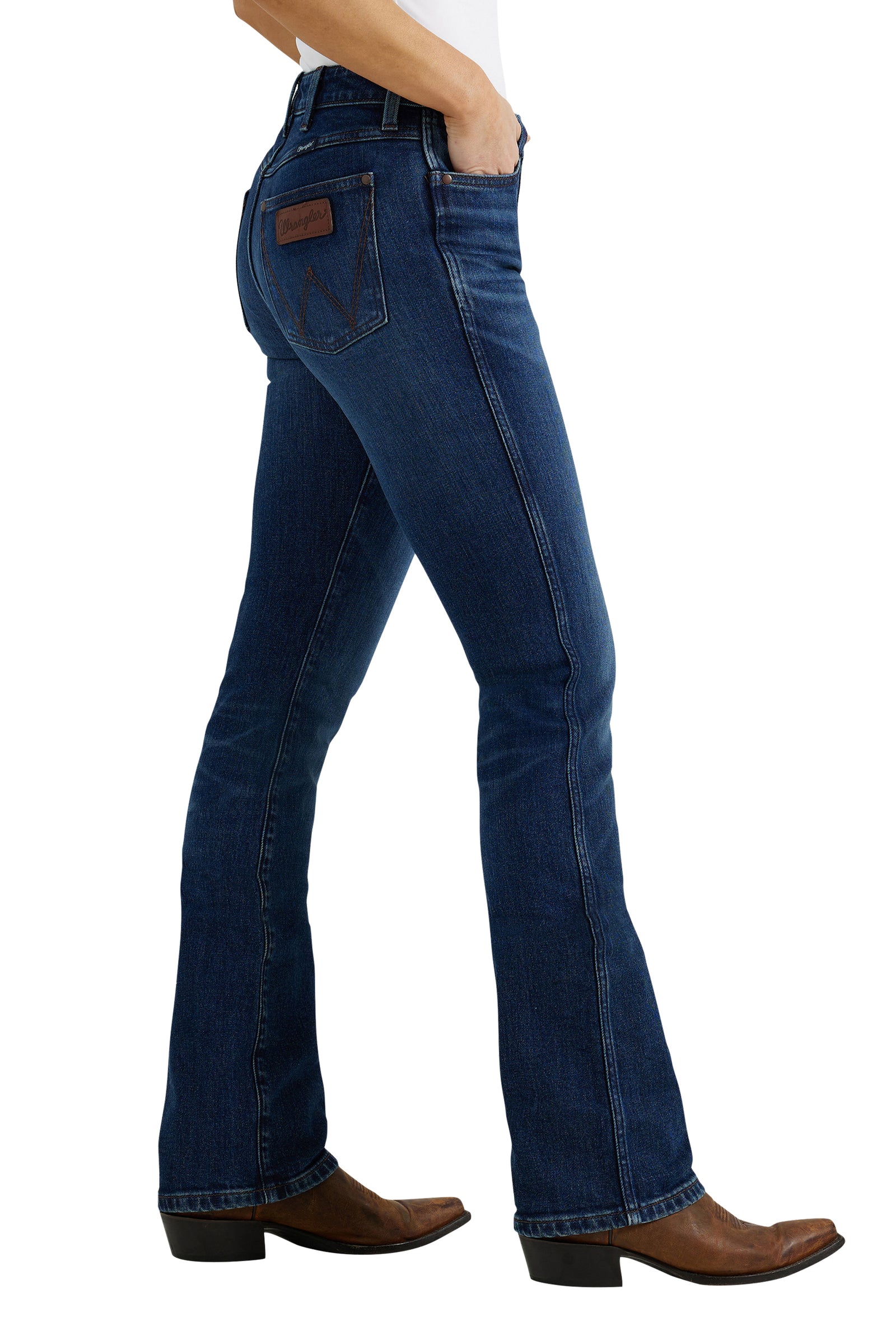 A woman wearing Wrangler Women's High Rise Boot Cut Jeans and brown boots, standing with one hand in pocket.