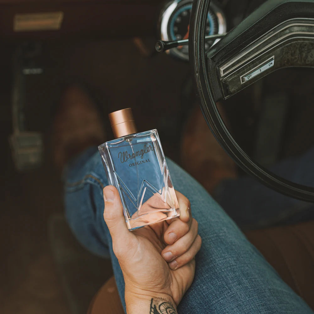A person in a car holds a bottle of Wrangler Original denim spray. The bottle is clear glass with a blue-gray geometric pattern, a metallic copper cap, and the Wrangler logo is printed on the front.